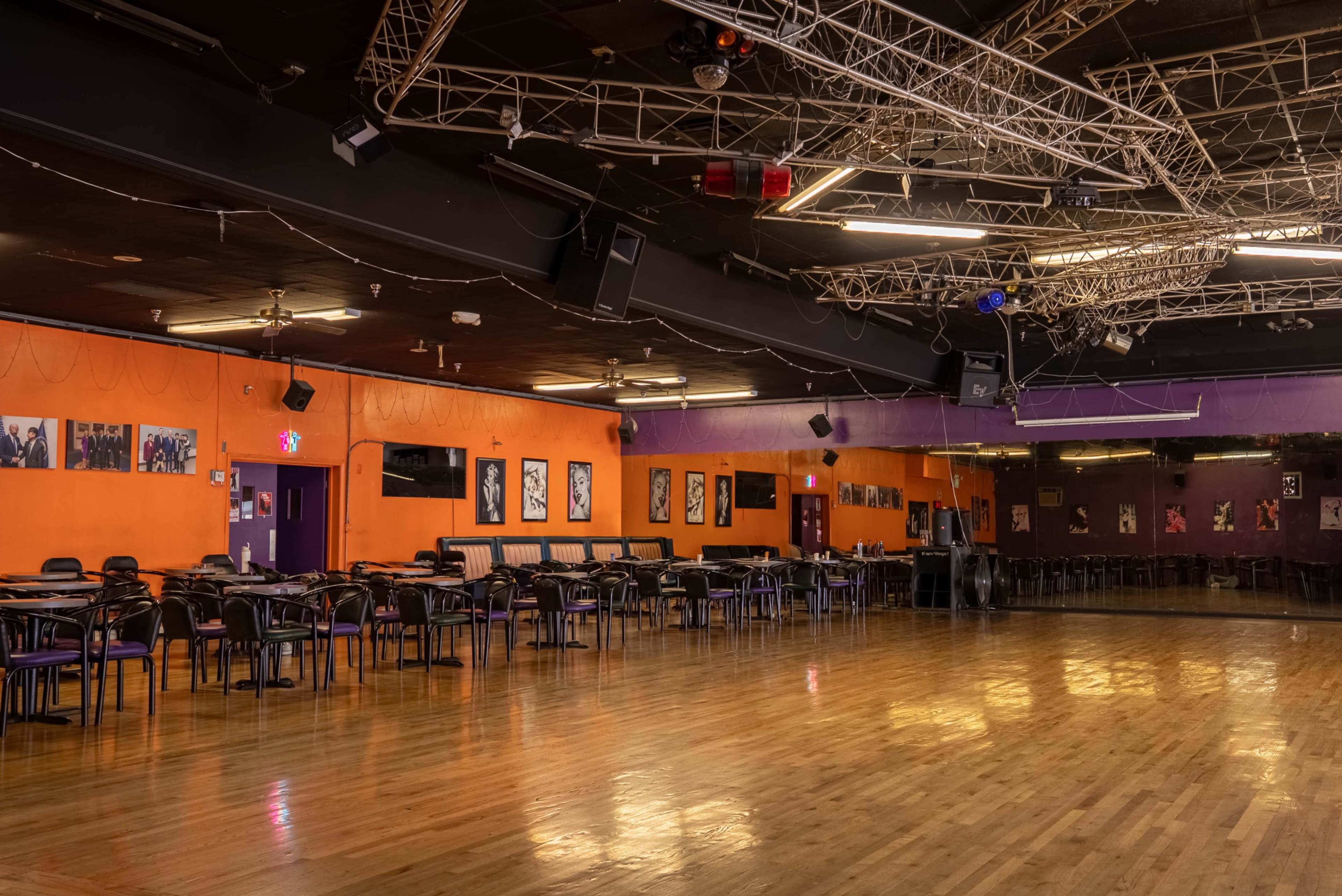 The image shows an empty dance hall with black and orange walls, wooden flooring, and rows of tables and chairs lined against the perimeter.