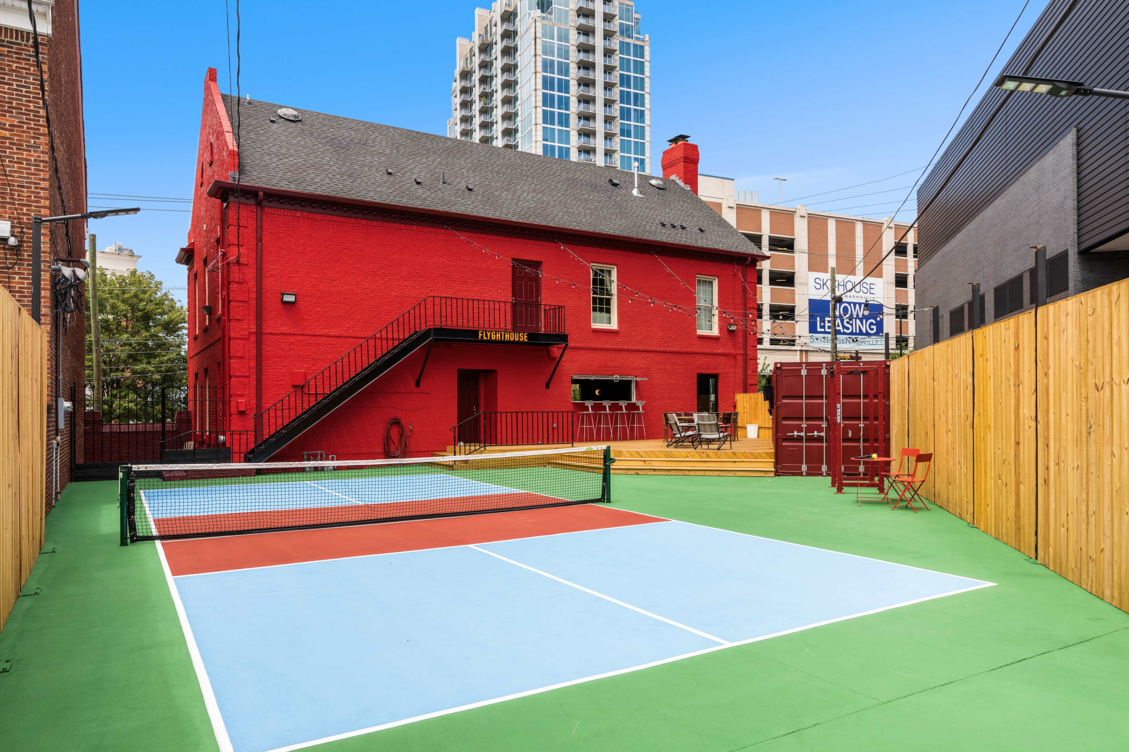 A red building with a staircase, adjacent to a green multi-use sports court featuring two distinct playing areas, set against a backdrop of tall buildings.