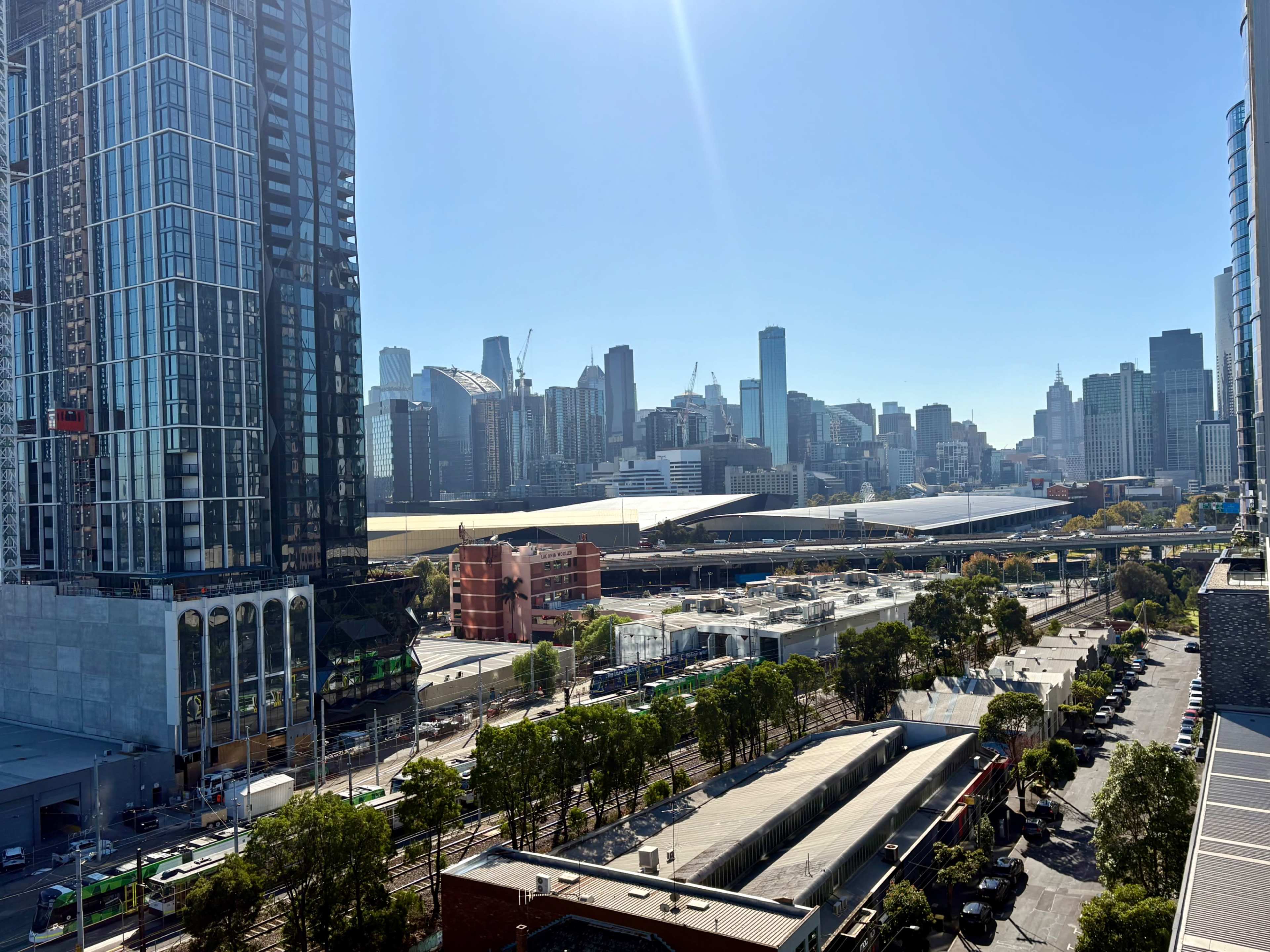 The image shows a view of a city skyline with tall buildings, a railway area, and a clear blue sky.
