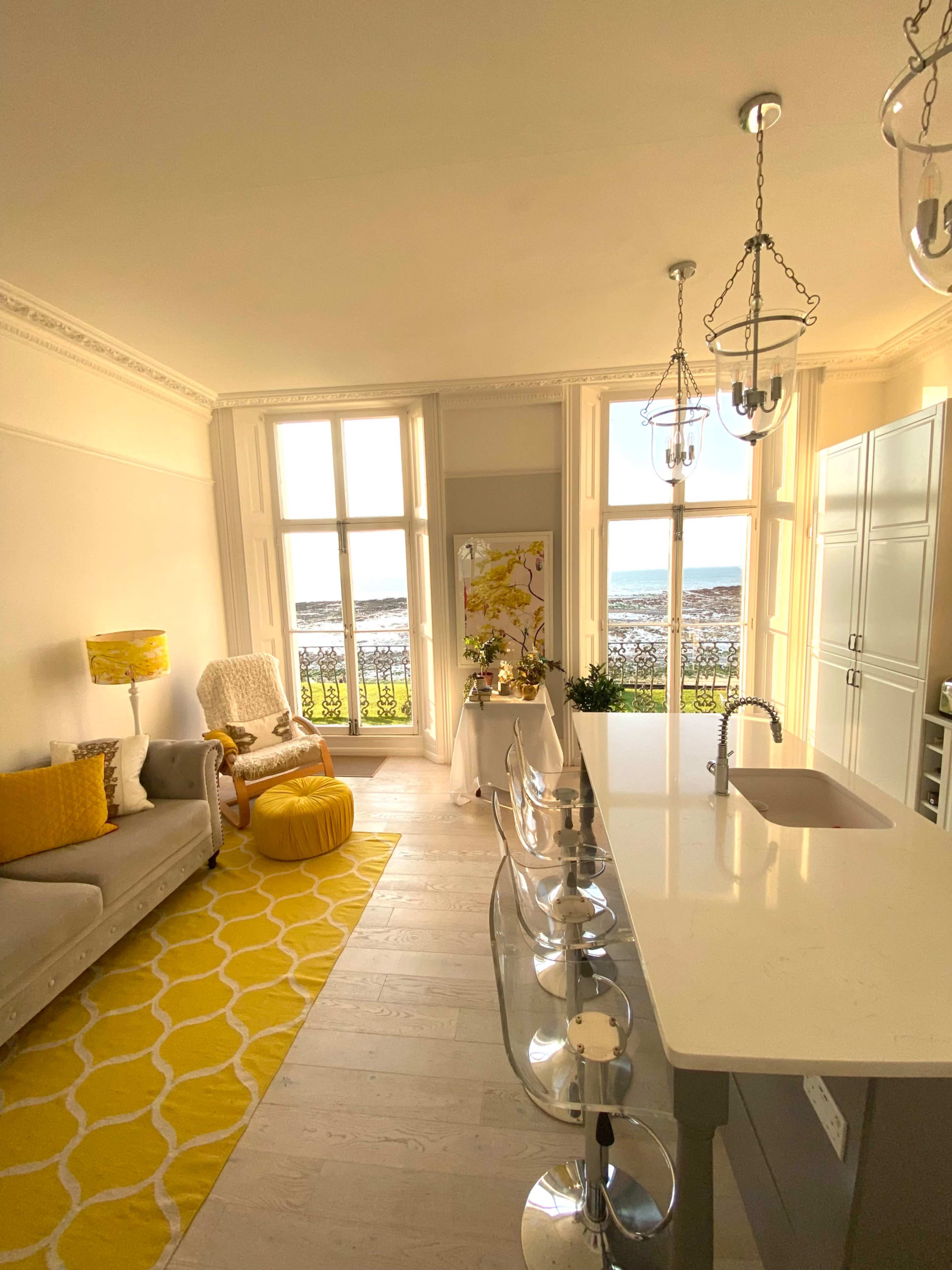 The image shows a bright kitchen and living area with large windows overlooking a body of water, featuring modern furnishings and a yellow color scheme.