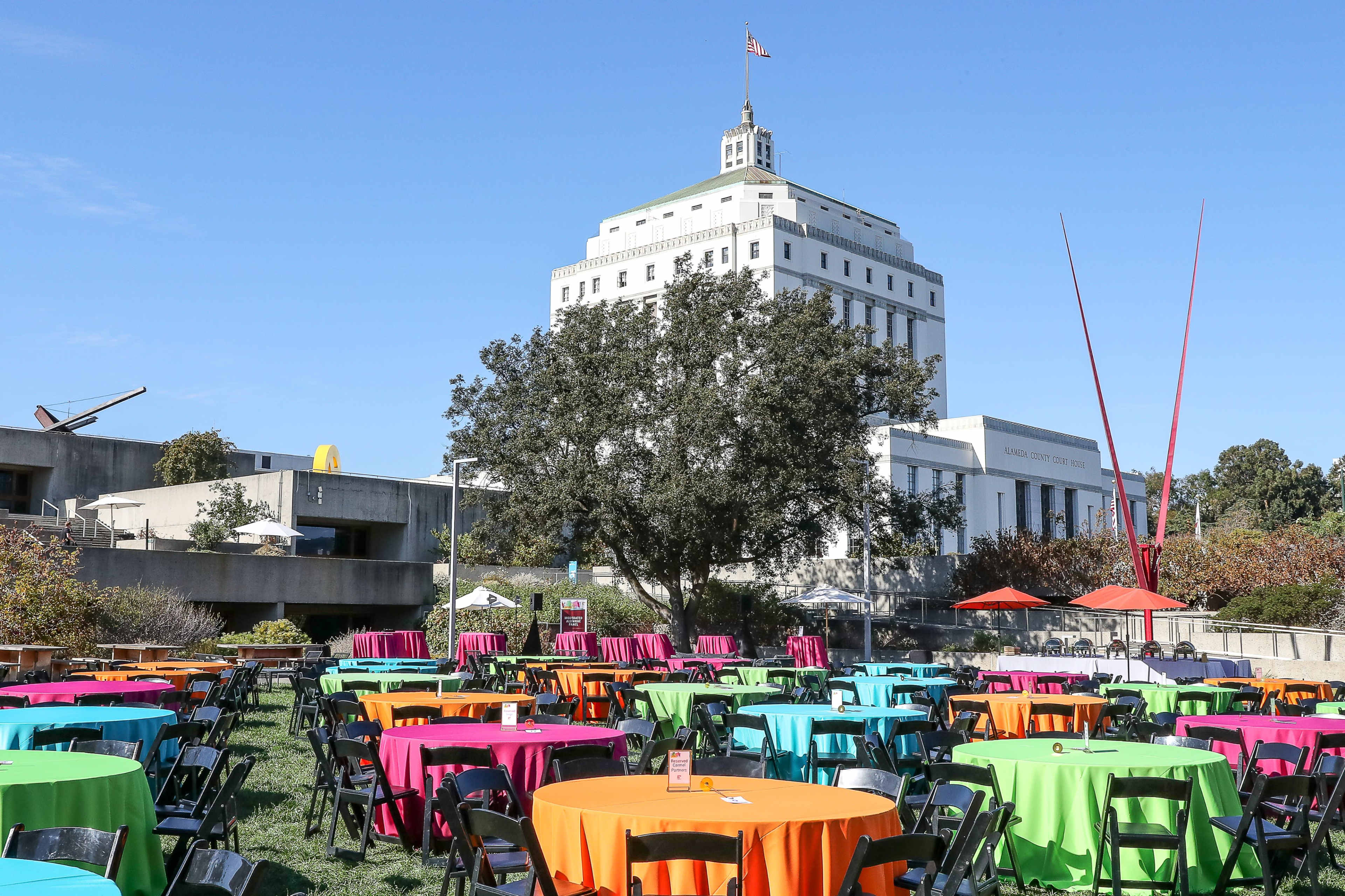 The scene features a vibrant outdoor event with colorful tables set up in front of a distinctive modern building under a clear blue sky.