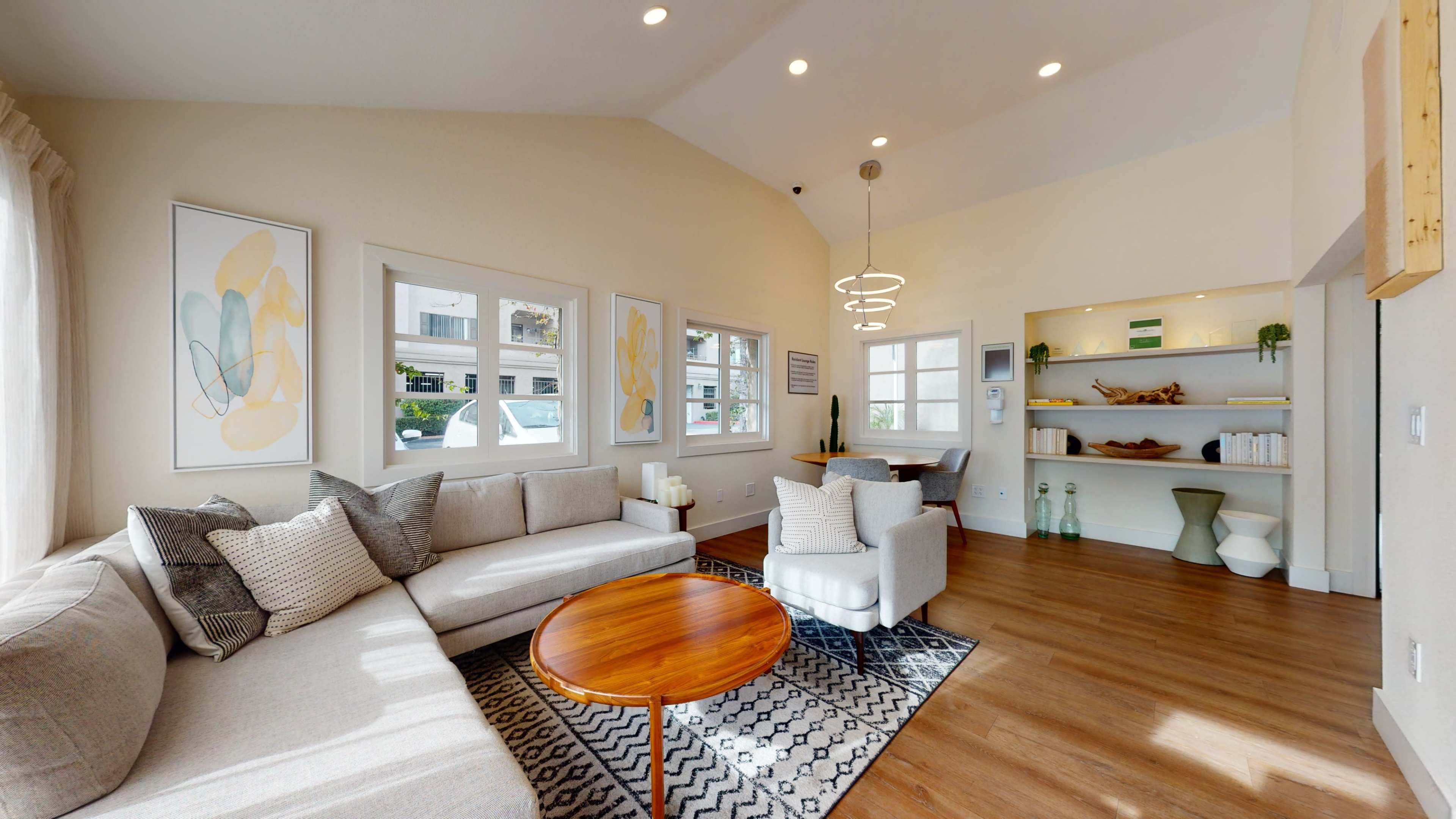 A modern living room with a light-colored sectional sofa, a circular wooden coffee table, and large windows allowing natural light, complemented by minimalist decor and wooden accents.