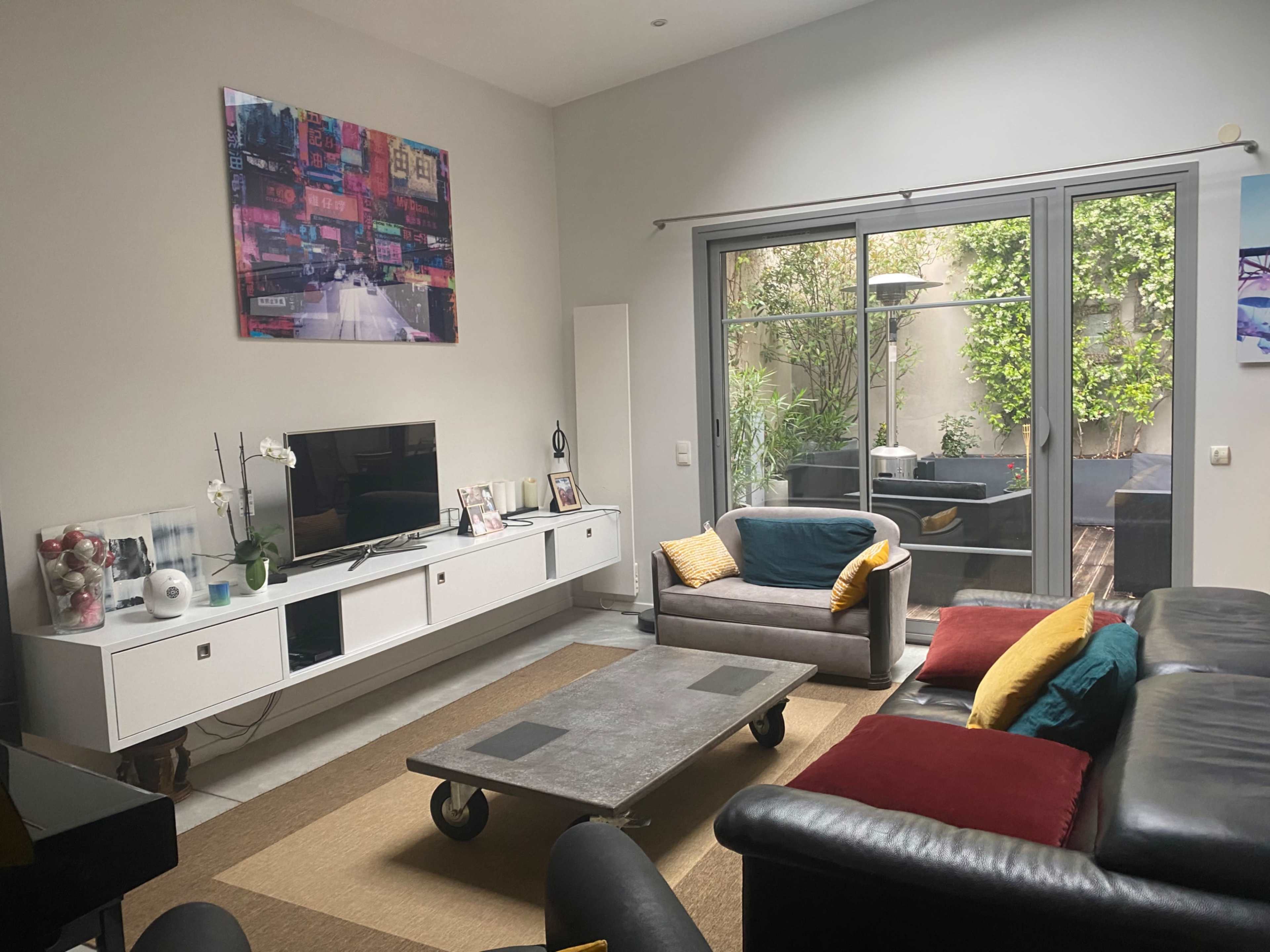A modern living room features a large TV on a white media console, a coffee table in the center, and seating arrangements with colorful cushions, overlooking a small outdoor space with greenery.