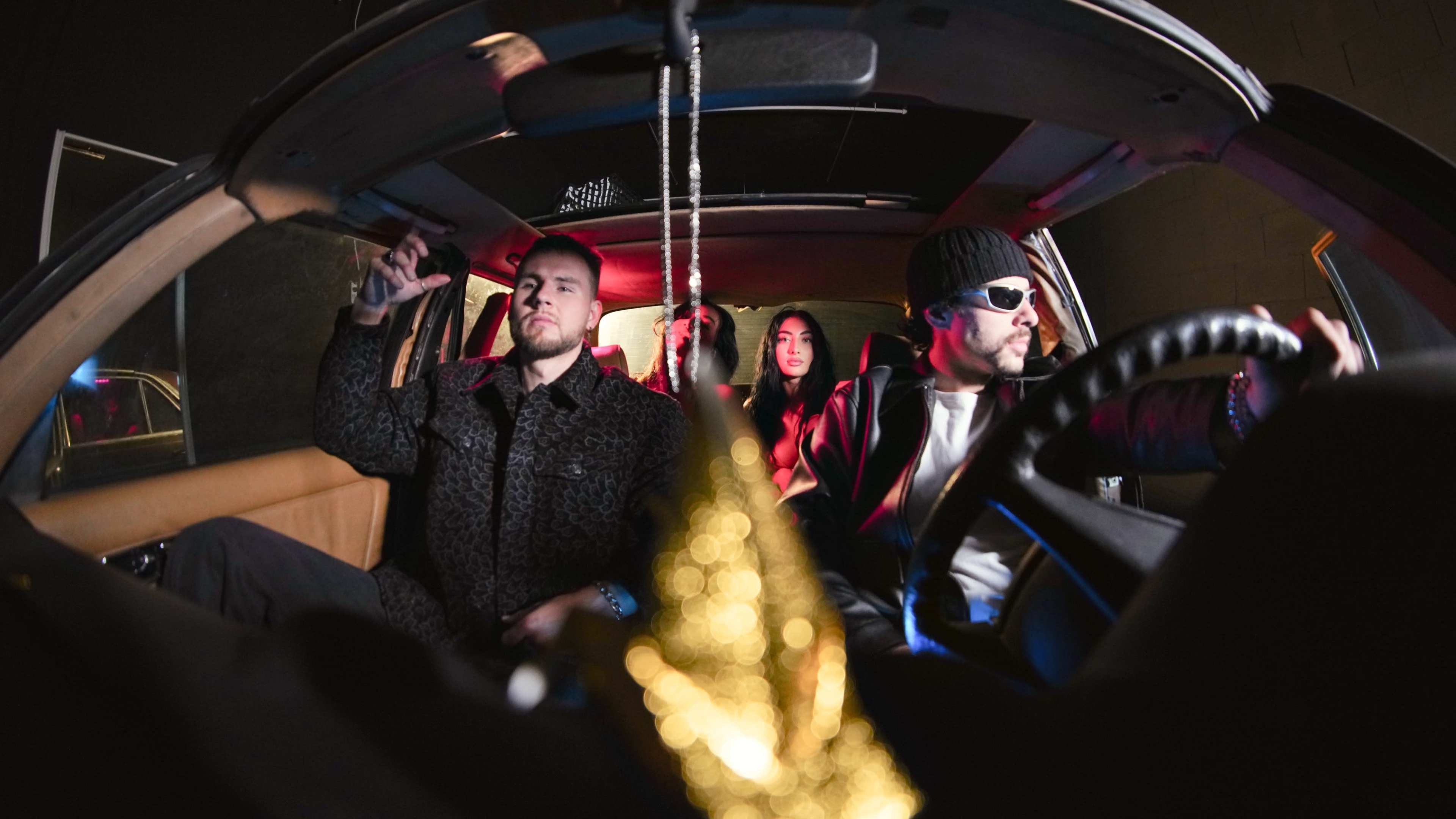 A group of four people sits inside a car, with one person in the front passenger seat gesturing and the driver wearing sunglasses.