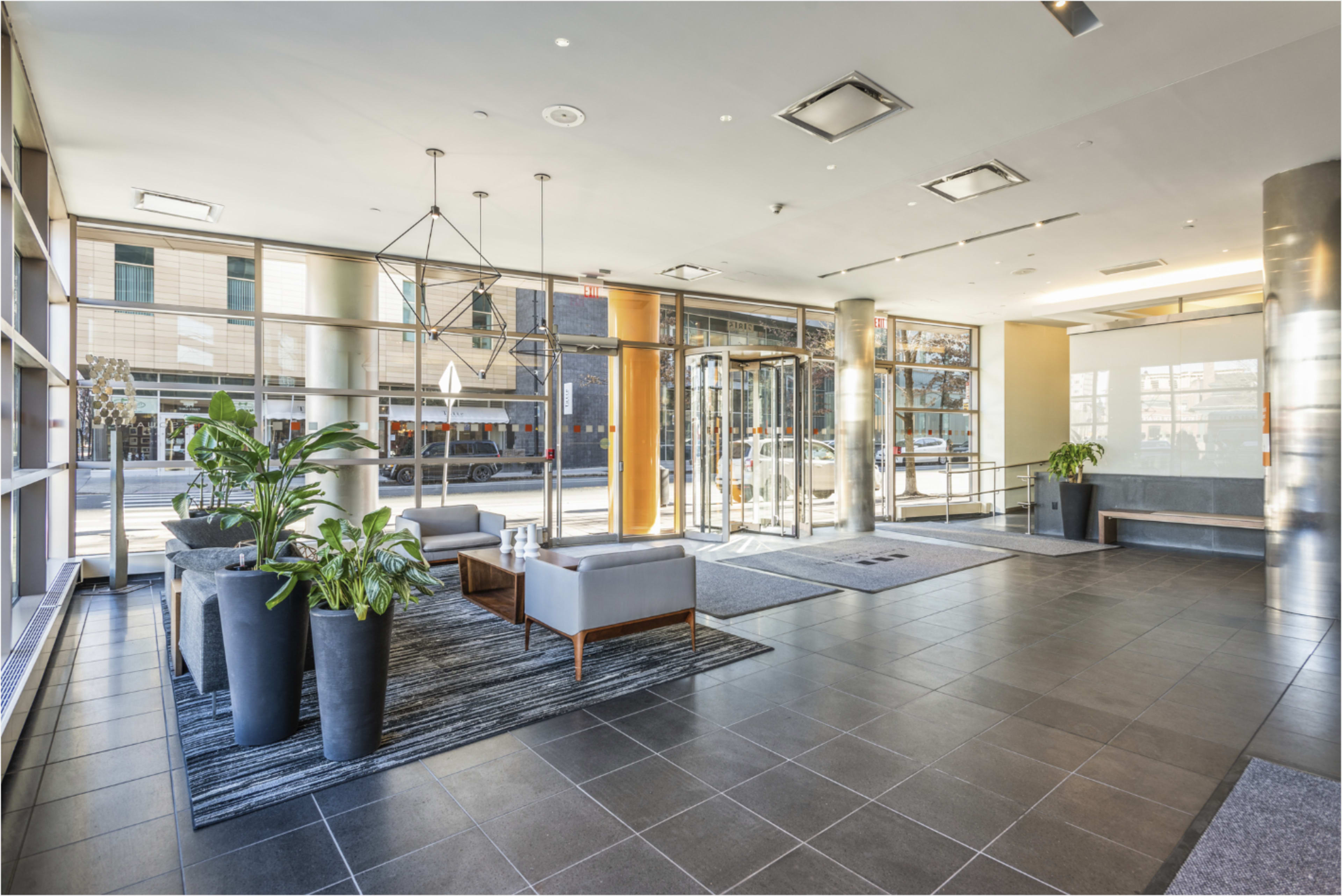The image shows a modern building lobby featuring a seating area with two gray couches, potted plants, and large glass doors leading to the outside.