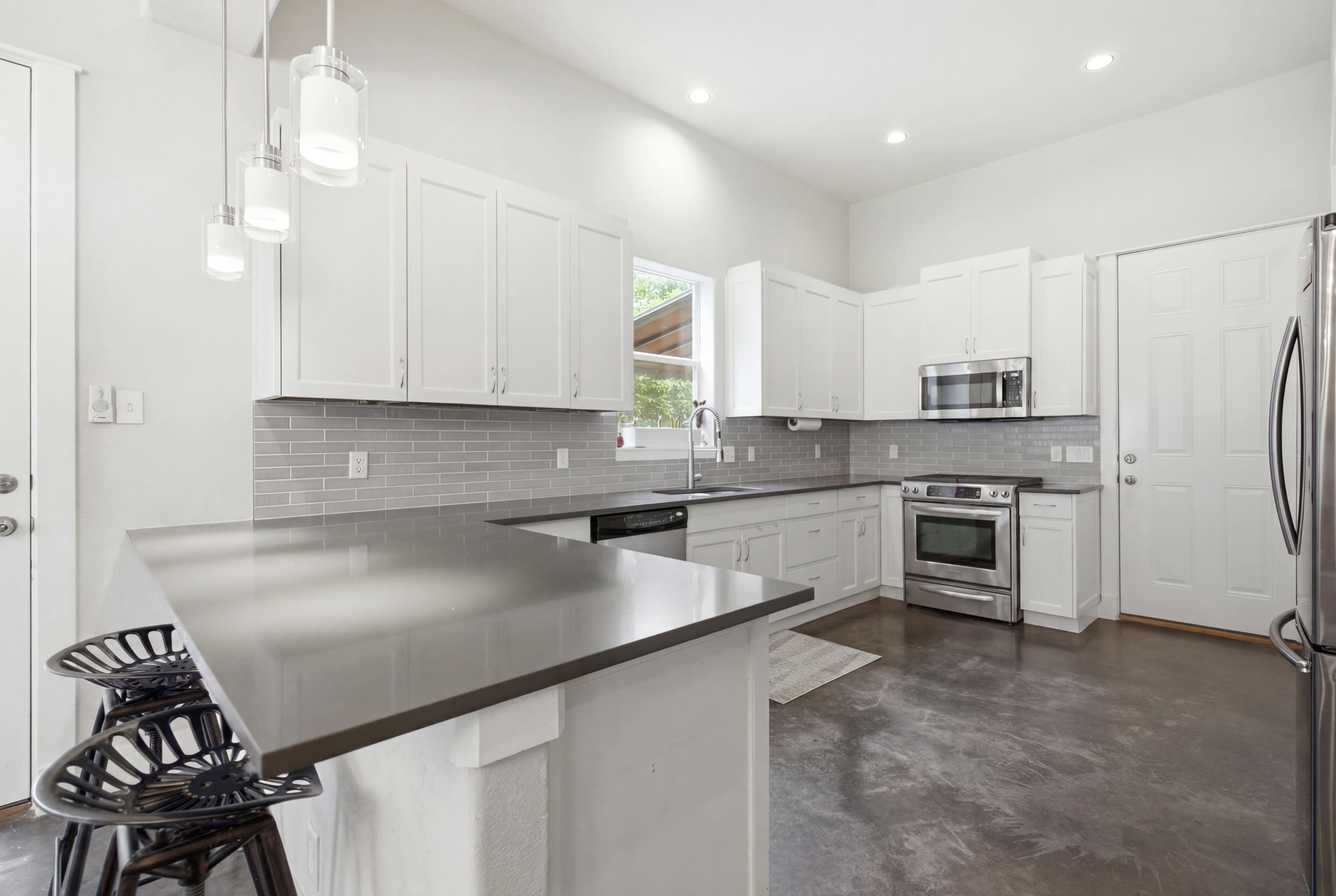 The image depicts a modern kitchen with white cabinetry, stainless steel appliances, and a concrete floor, featuring a large island with black bar stools.