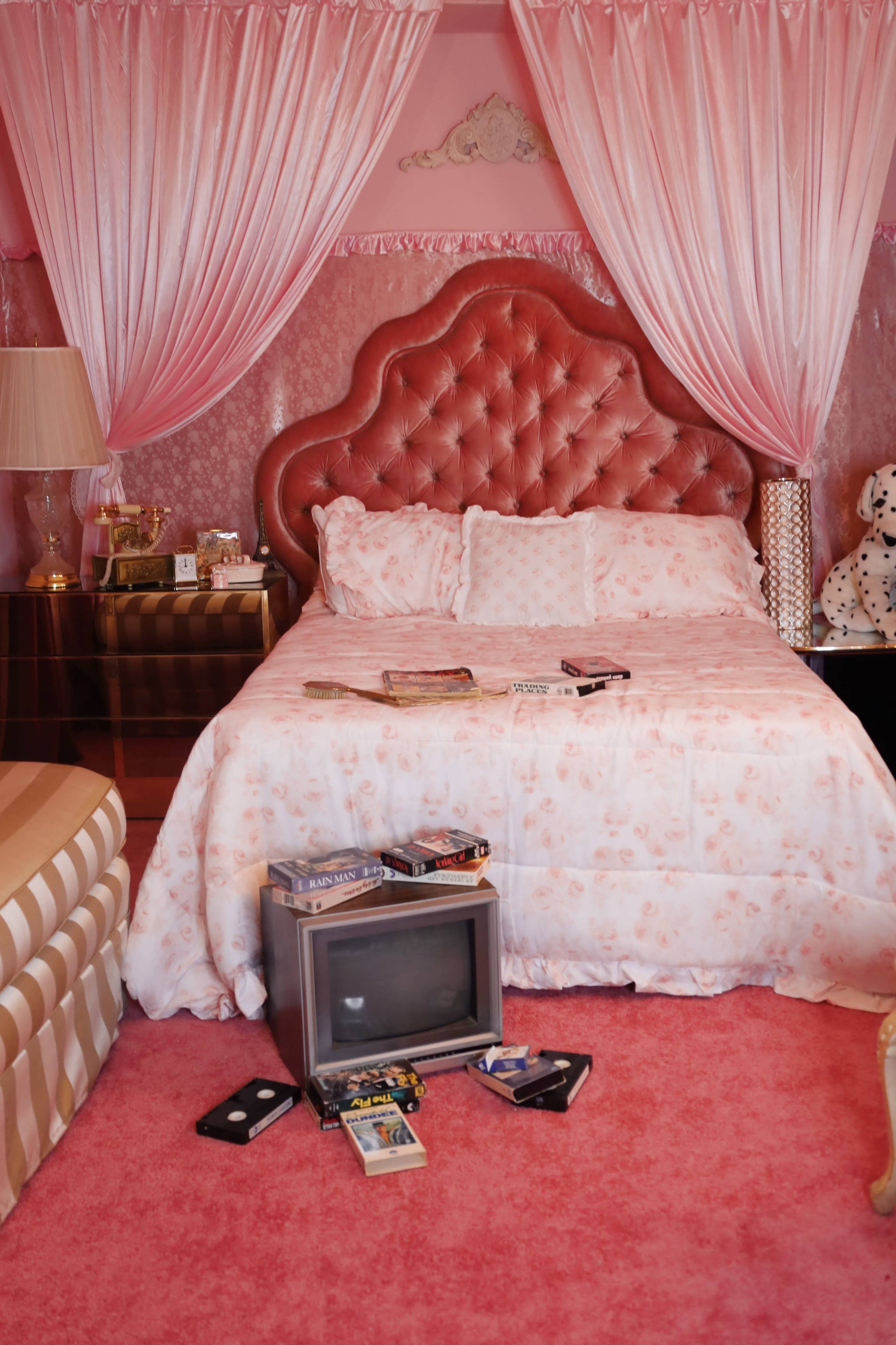 The scene features a pink-themed bedroom with a tufted headboard bed, a retro television on the floor, and various video cassettes scattered around.