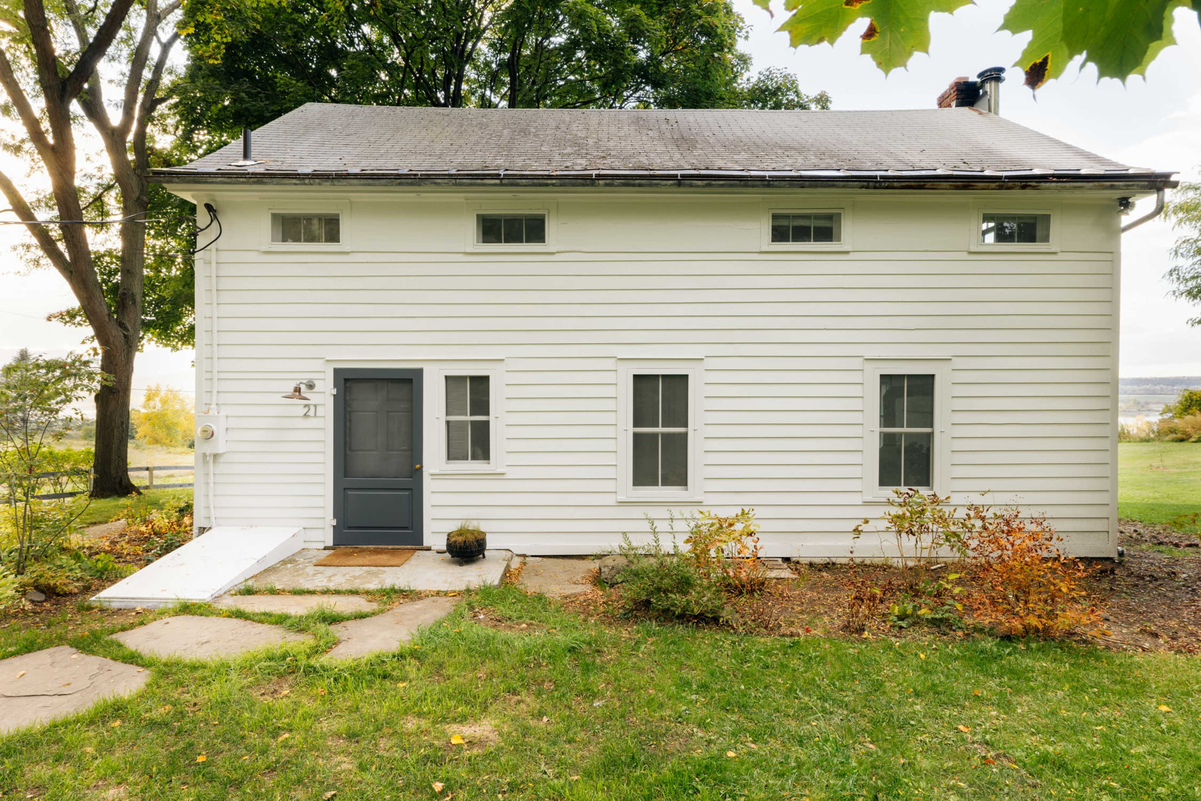A white, two-story house with a dark gray front door and several small windows, set in a grassy area with a concrete path leading up to it.