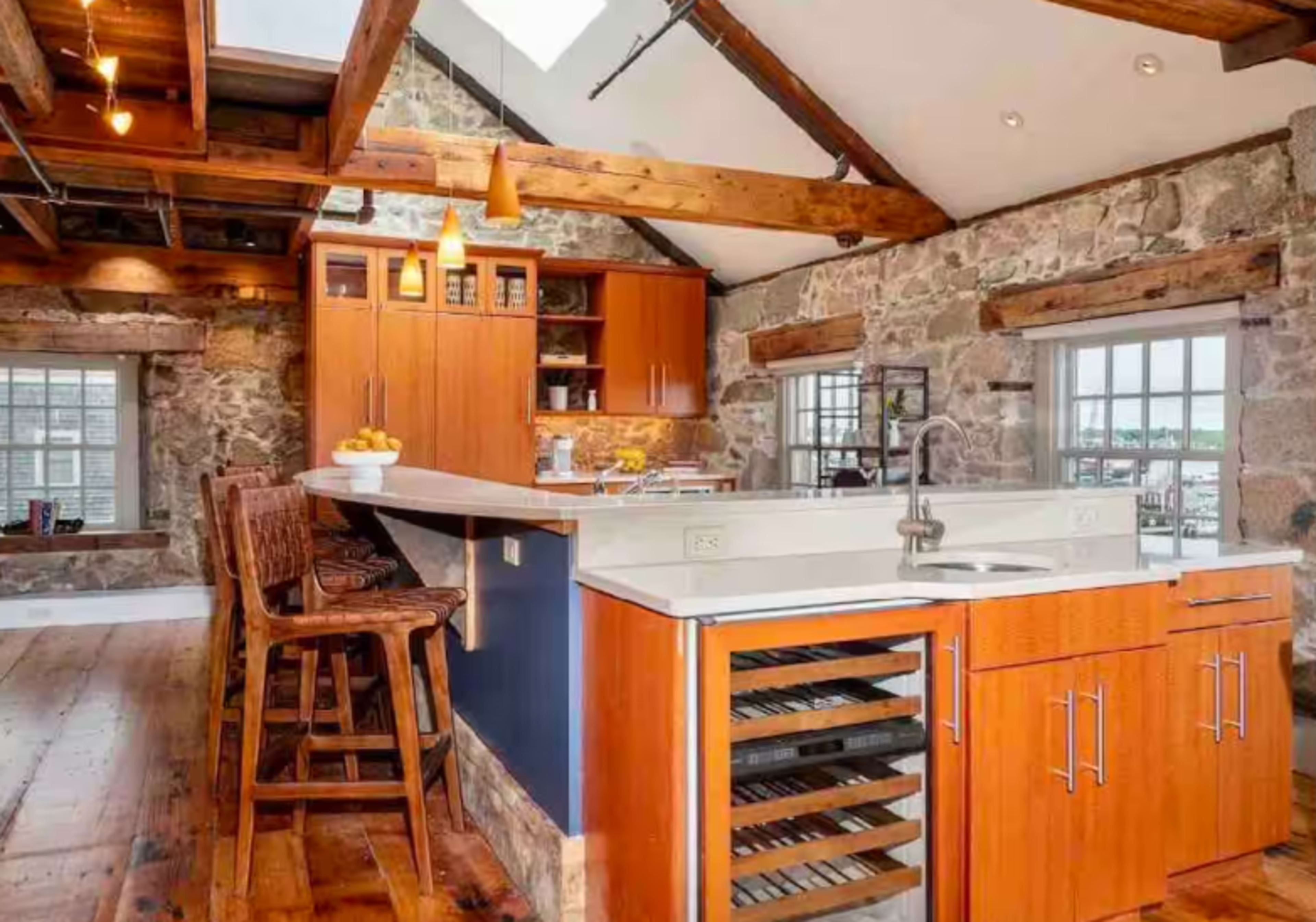 The image shows a rustic kitchen featuring stone walls, wooden beams, and an island with bar stools, along with modern appliances and a wine cooler.