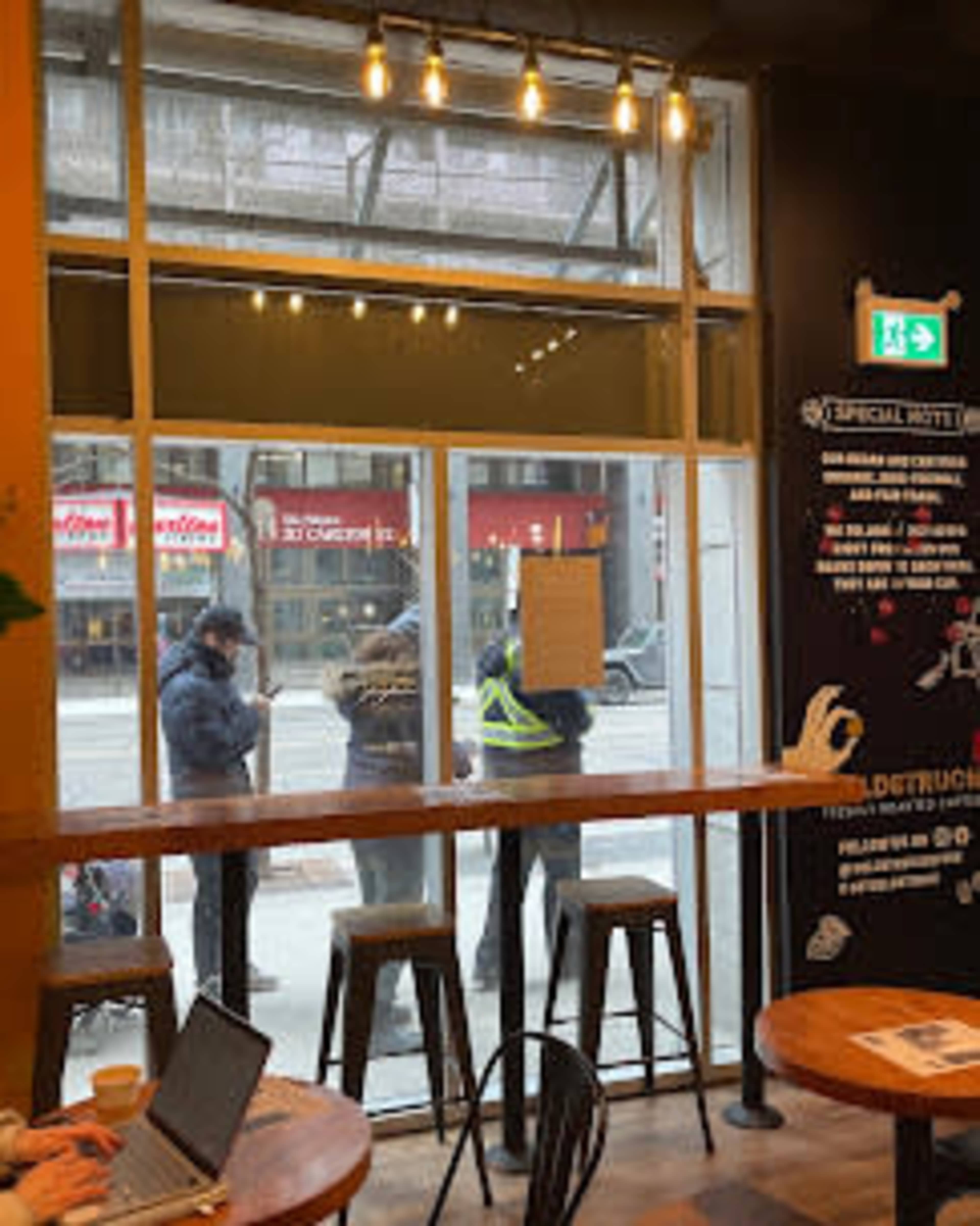 The image shows a coffee shop interior with a bar-like table and stools, while several people are gathered outside near the large window.