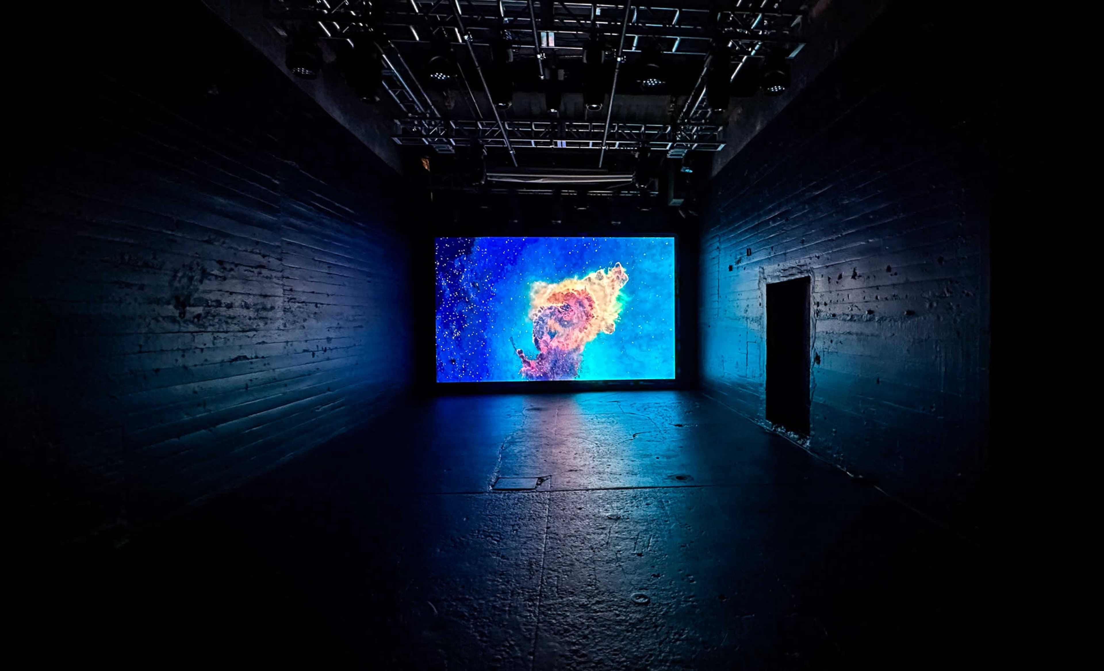 A large screen displaying colorful visuals is positioned in a dark, industrial-looking room with textured walls.