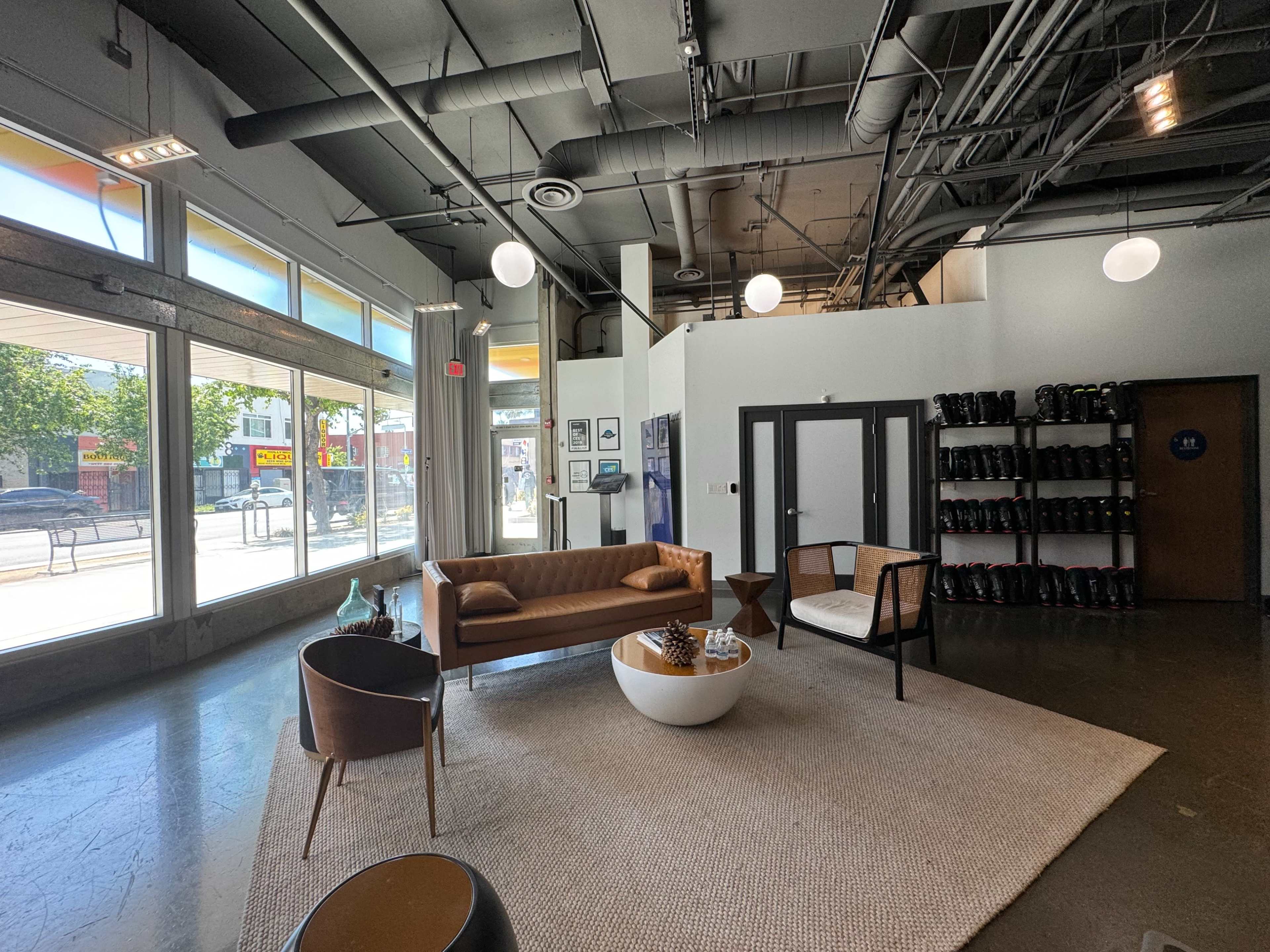 The interior of a modern waiting area features a brown leather sofa, a round white coffee table, and a black and white chair, surrounded by large windows and minimalist decor.