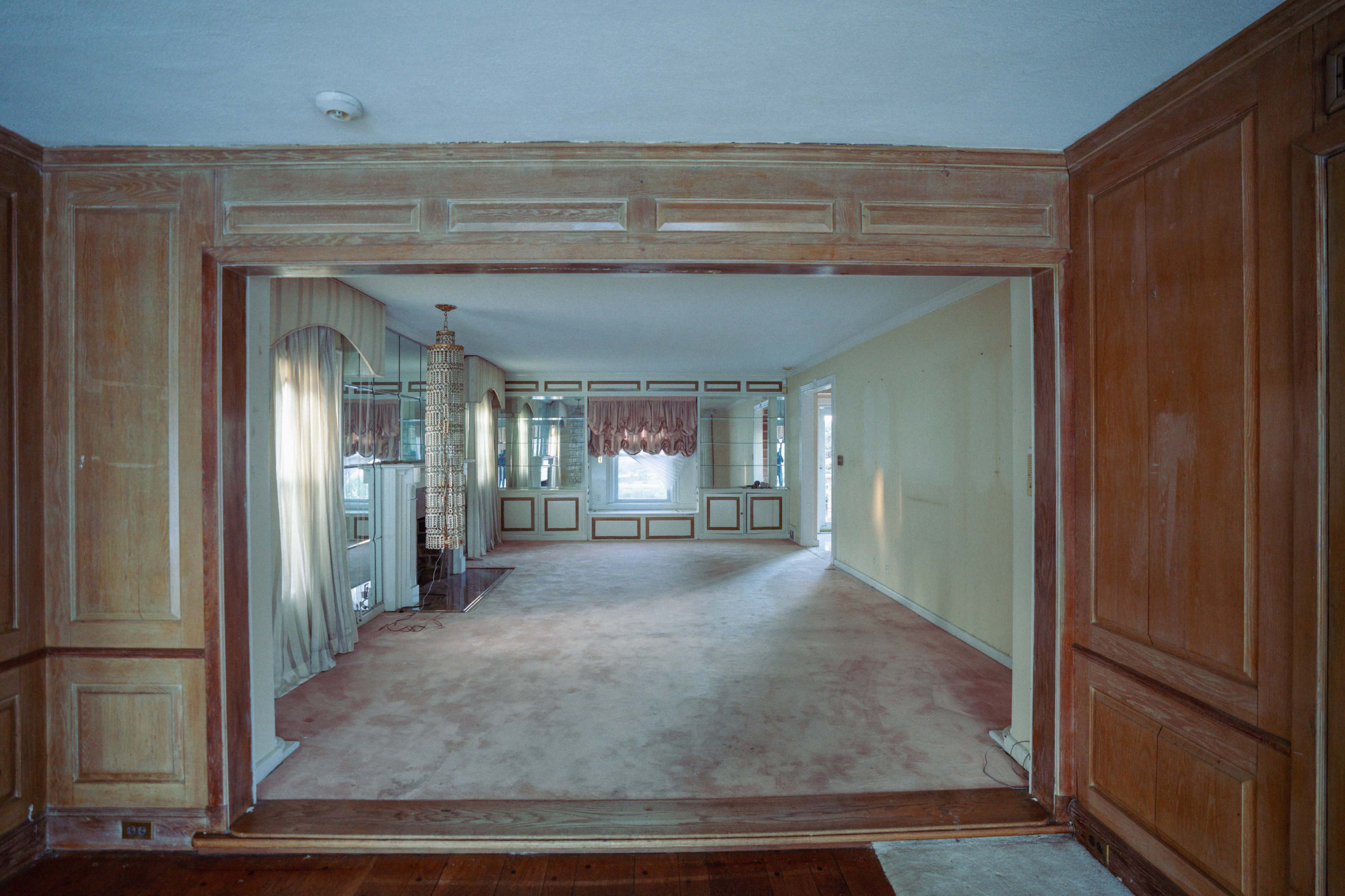 The image shows an empty, partially finished living room with wood paneling and beige carpeting, leading to a space with large windows and curtains.
