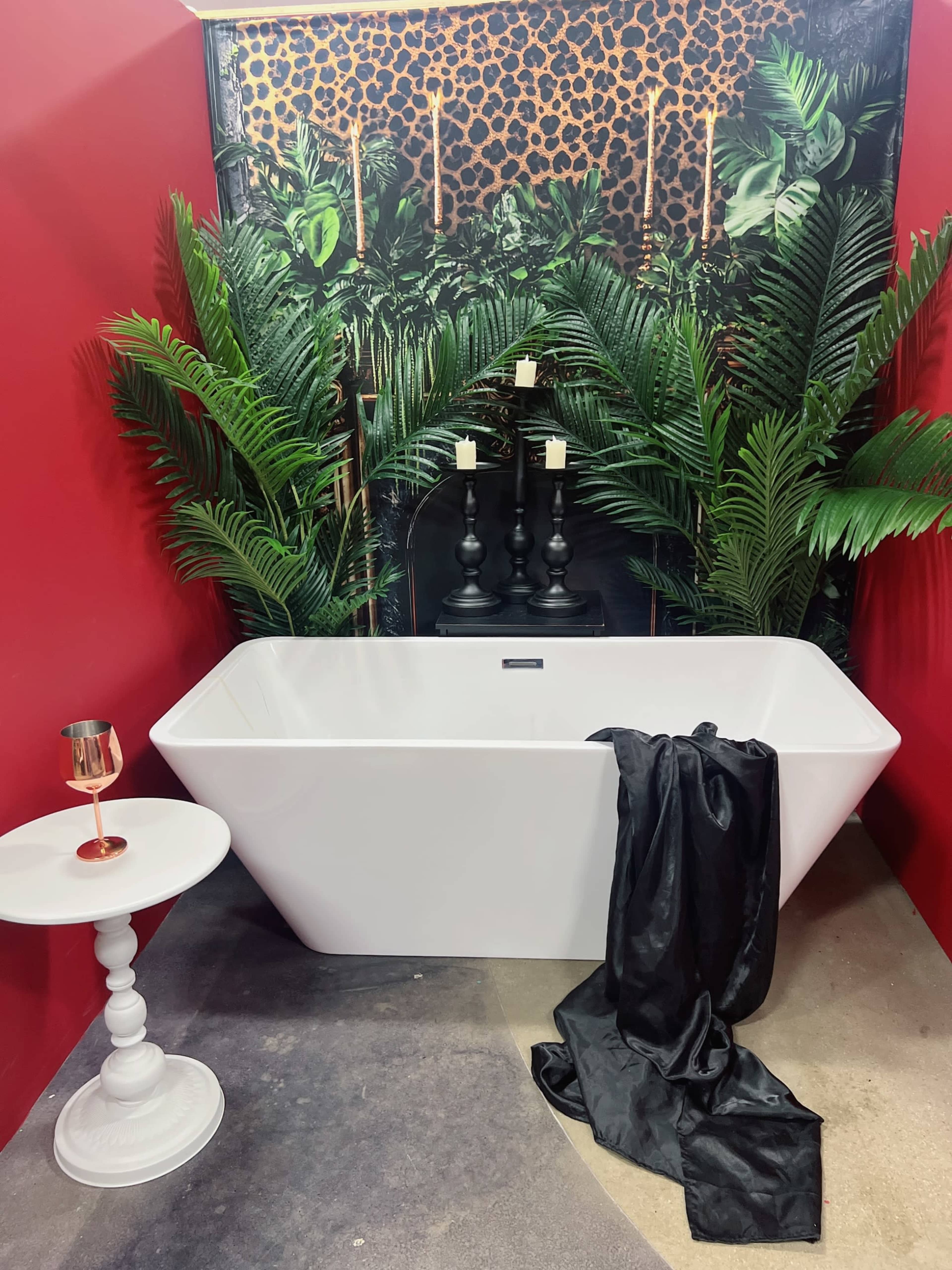 The image features a modern white freestanding bathtub positioned against a vibrant red wall adorned with tropical foliage and a leopard print backdrop.