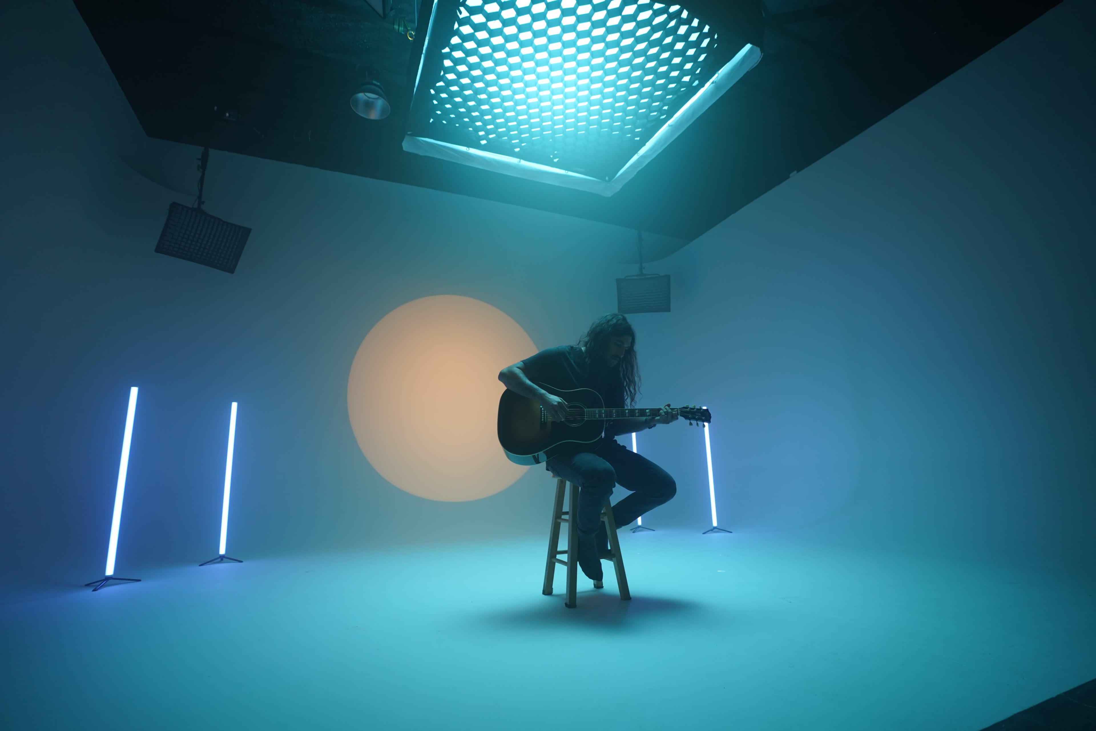 A musician plays an acoustic guitar while seated on a stool in a dimly lit studio with colored lighting and a large circular backdrop.