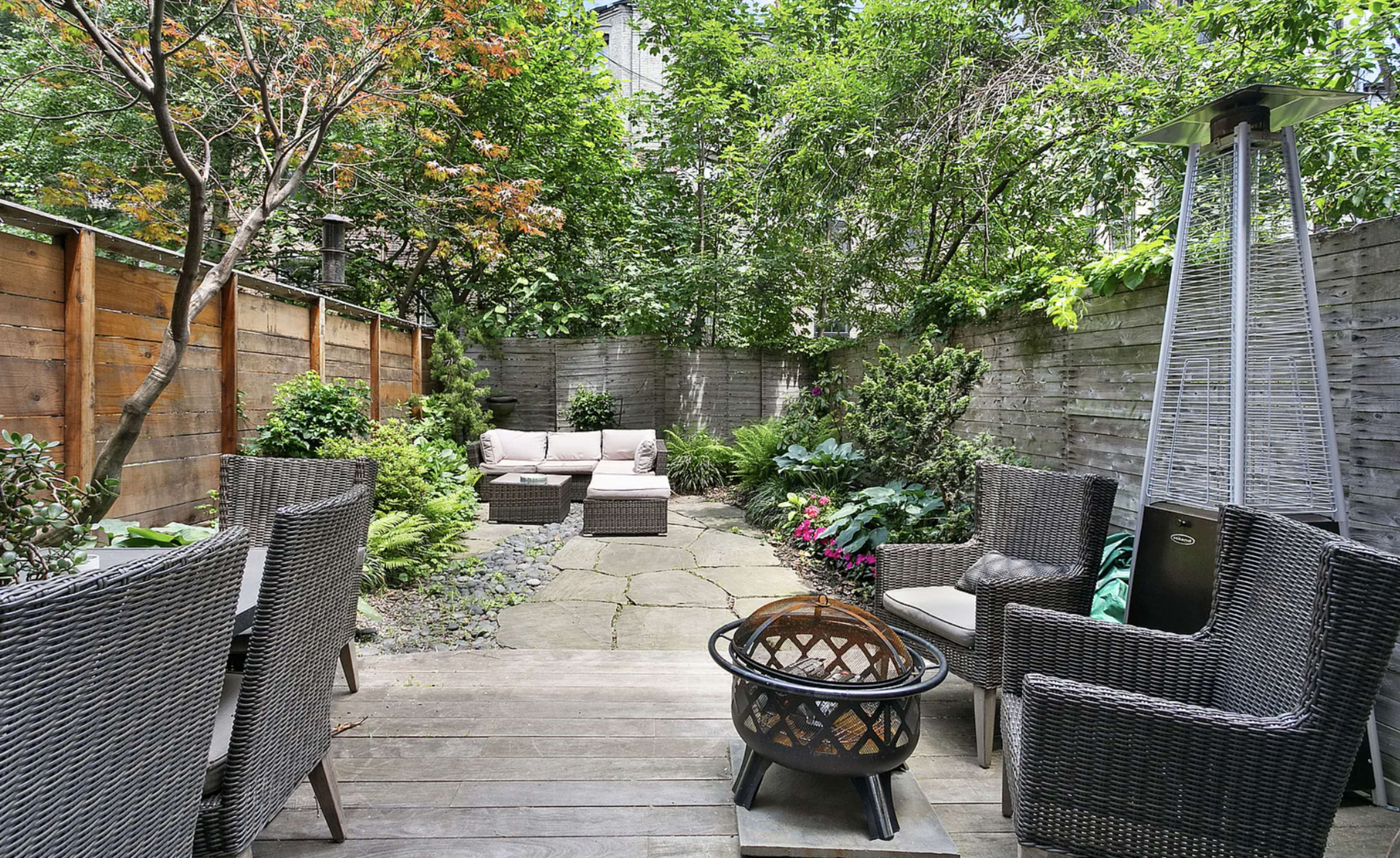 The image shows a landscaped backyard patio featuring a seating area with wicker furniture, a fire pit, and surrounding greenery.