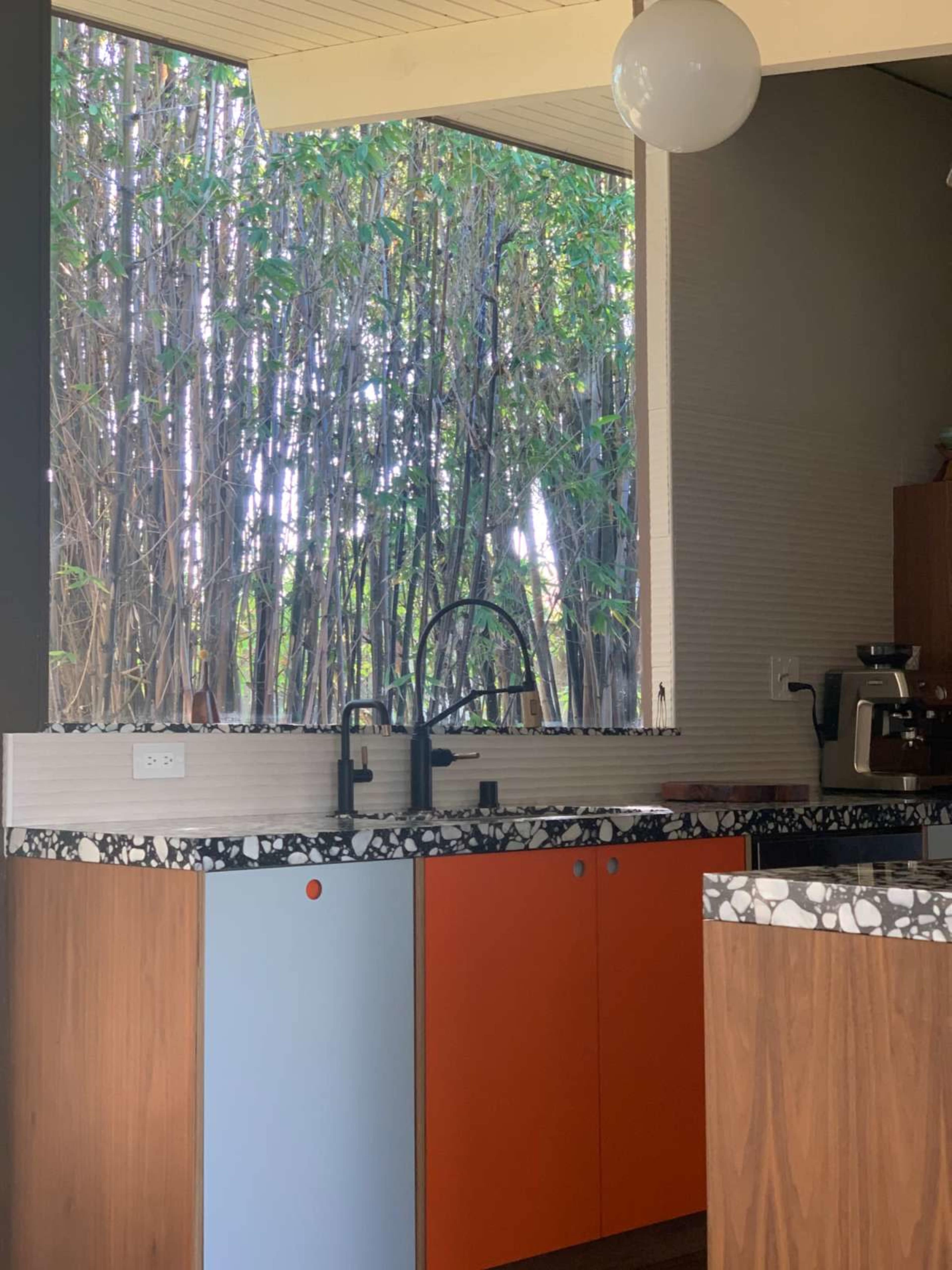 The image shows a modern kitchen with a mix of orange and blue cabinetry, a textured countertop, and a view of bamboo outside through a large window.
