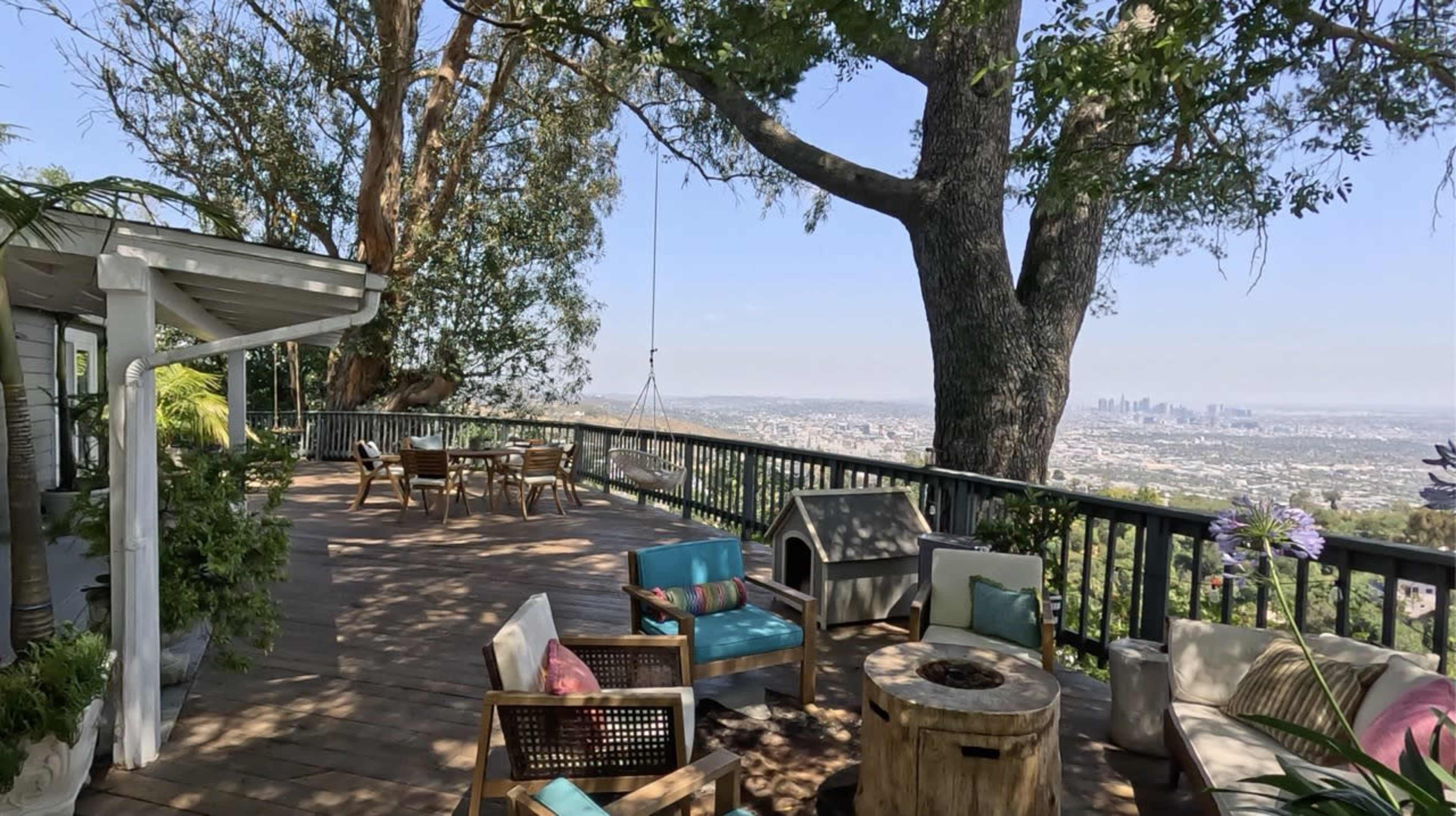 The image shows a spacious wooden deck overlooking a city skyline, furnished with outdoor seating and tables surrounded by trees.