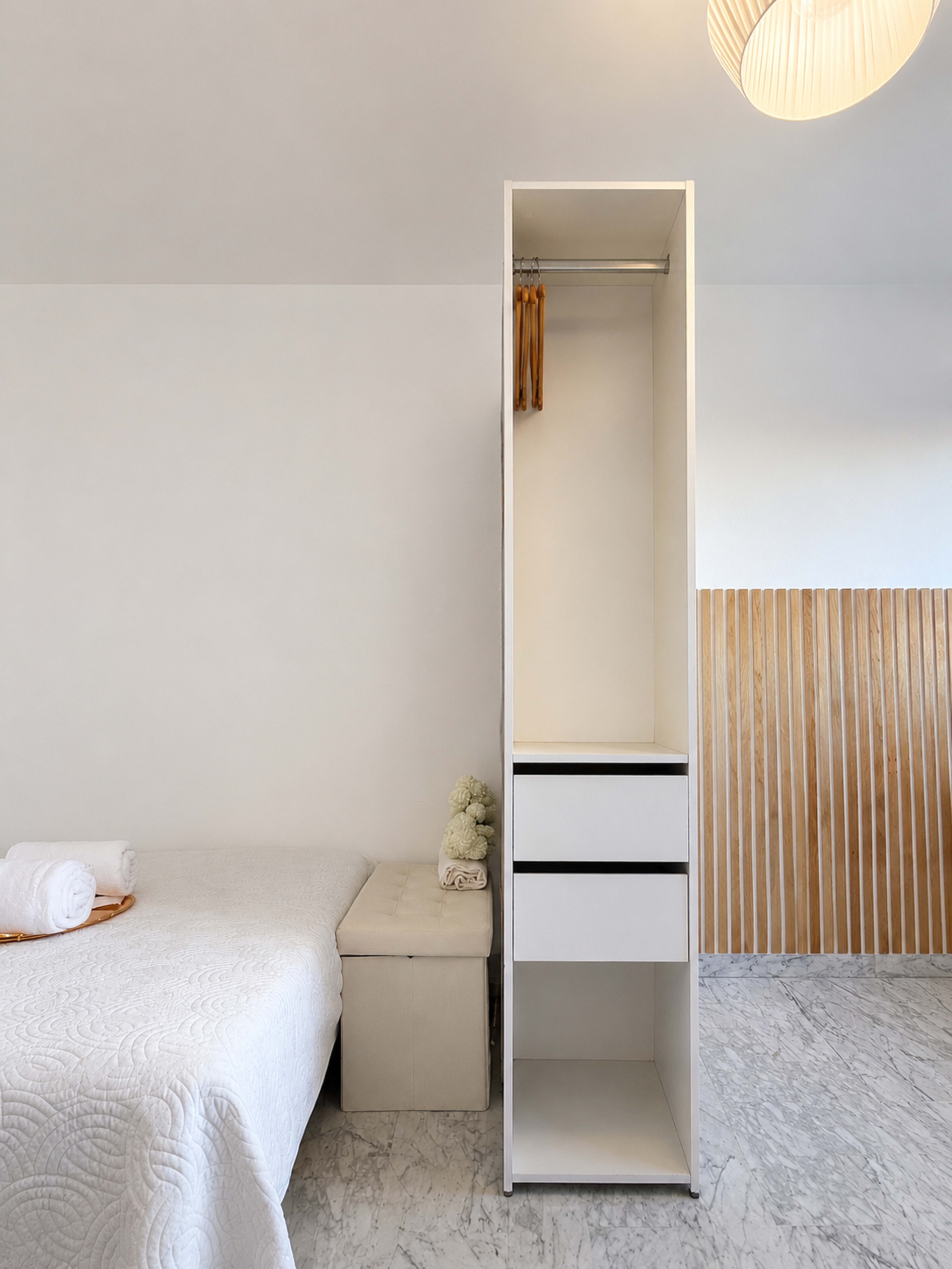 The image shows a simple bedroom with a bed, a light-colored storage bench, and a tall white wardrobe with hanging space and drawers.