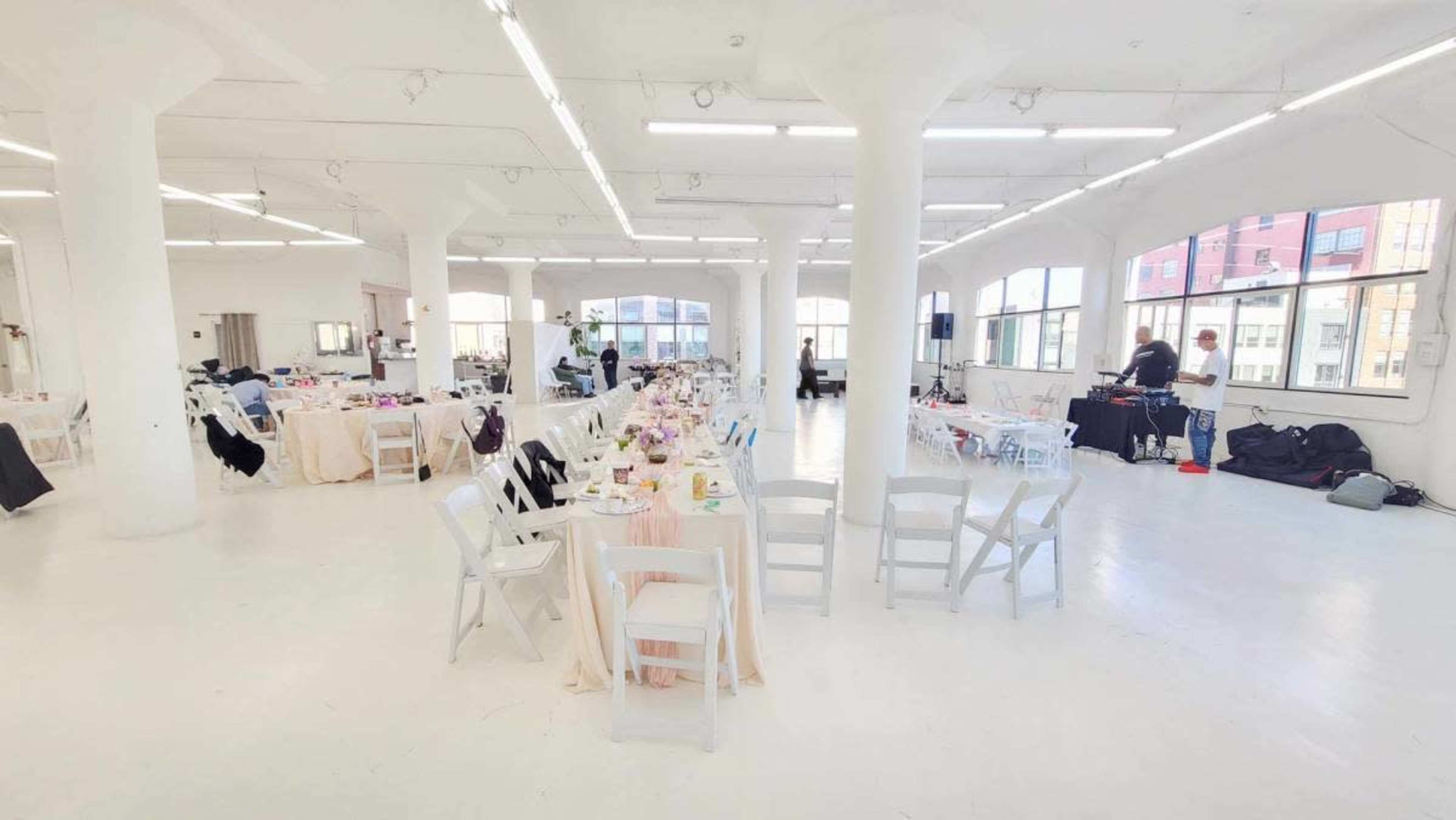 A spacious event hall features multiple tables set with light-colored tablecloths and chairs arranged throughout the room under bright overhead lighting.