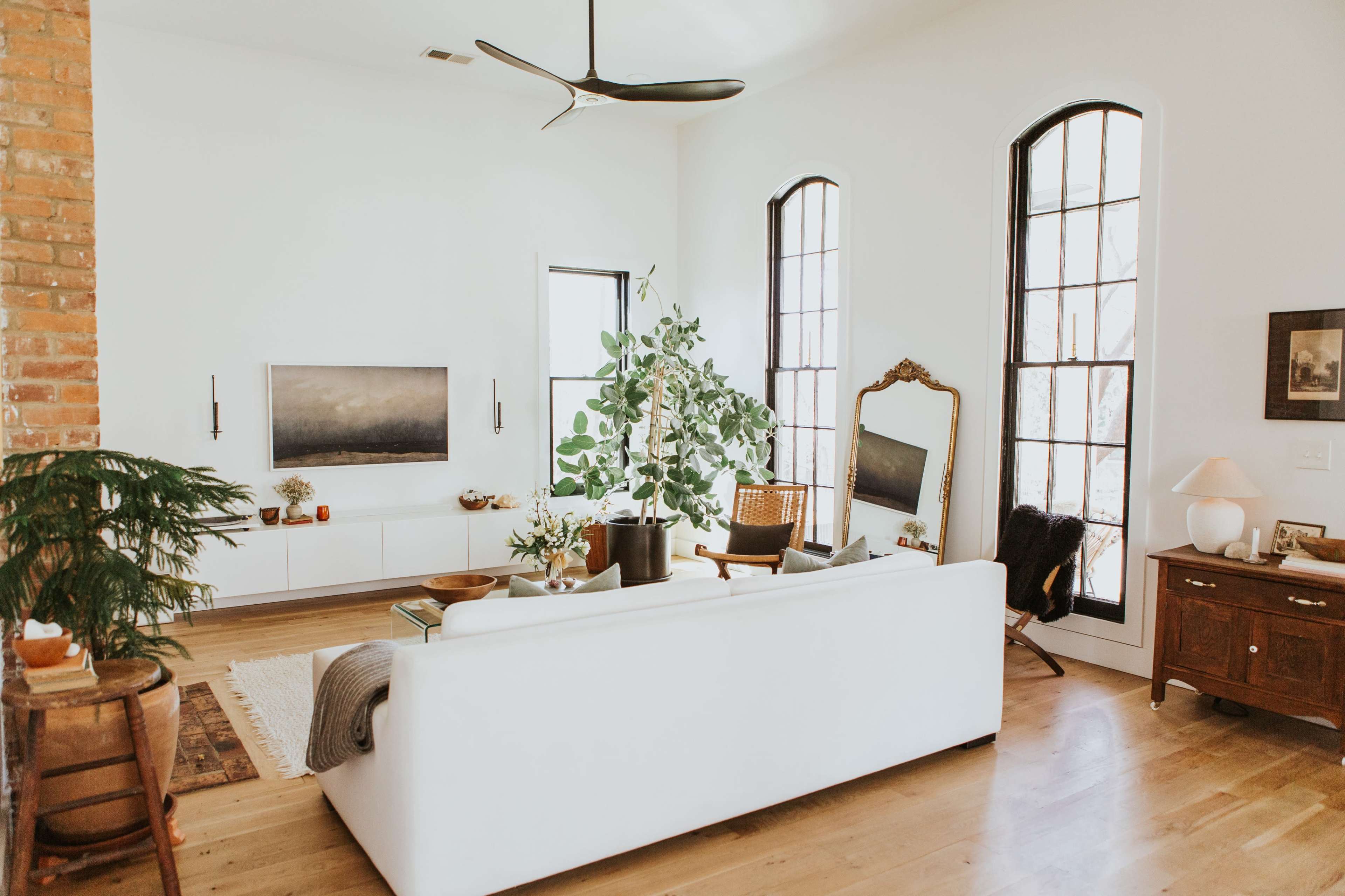 A bright living room features a white sofa, a leafy potted plant, and a large window, complemented by minimalist decor and wooden furniture.