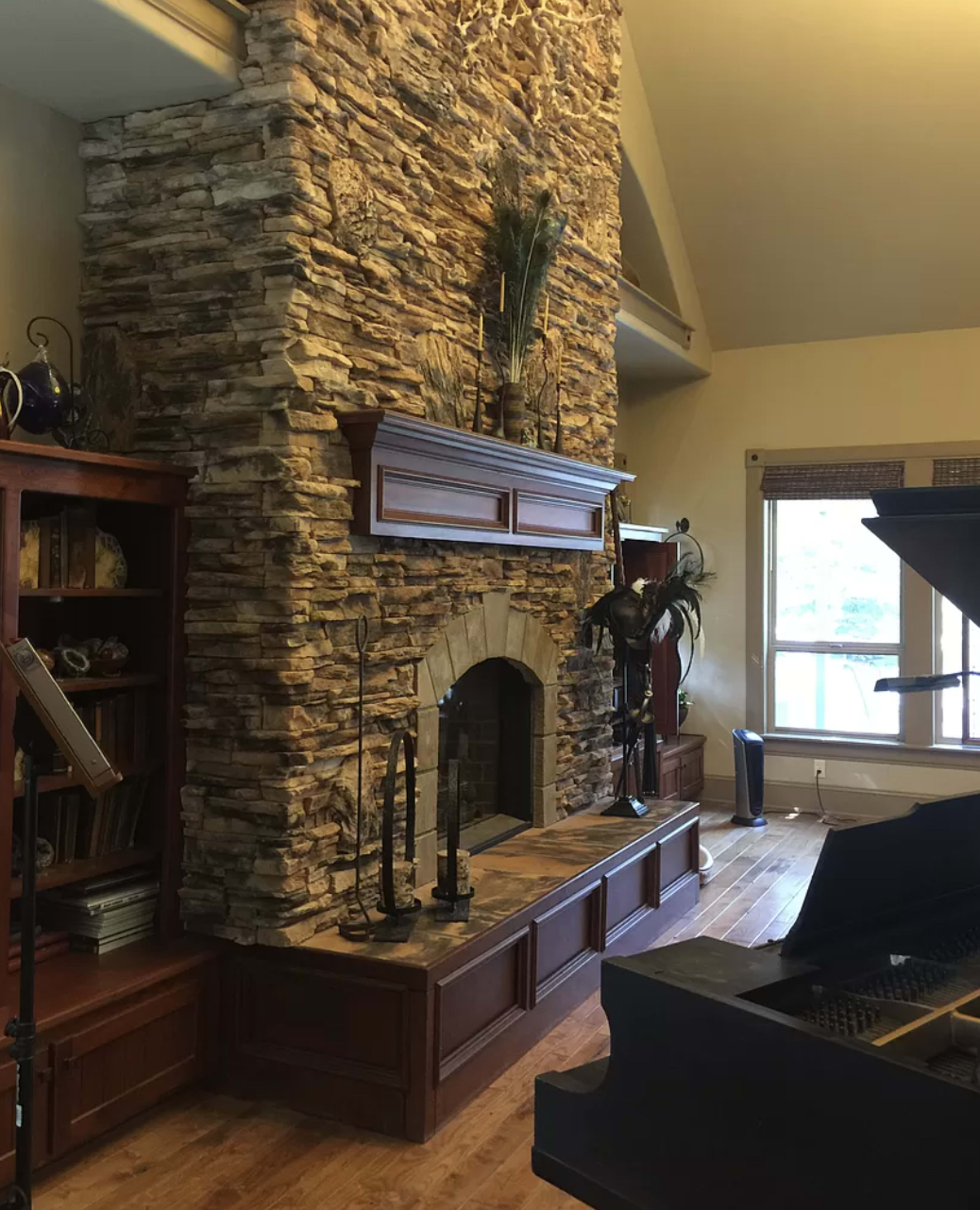 A living room featuring a large stone fireplace with a wooden mantel, surrounded by built-in shelves and a piano in one corner.