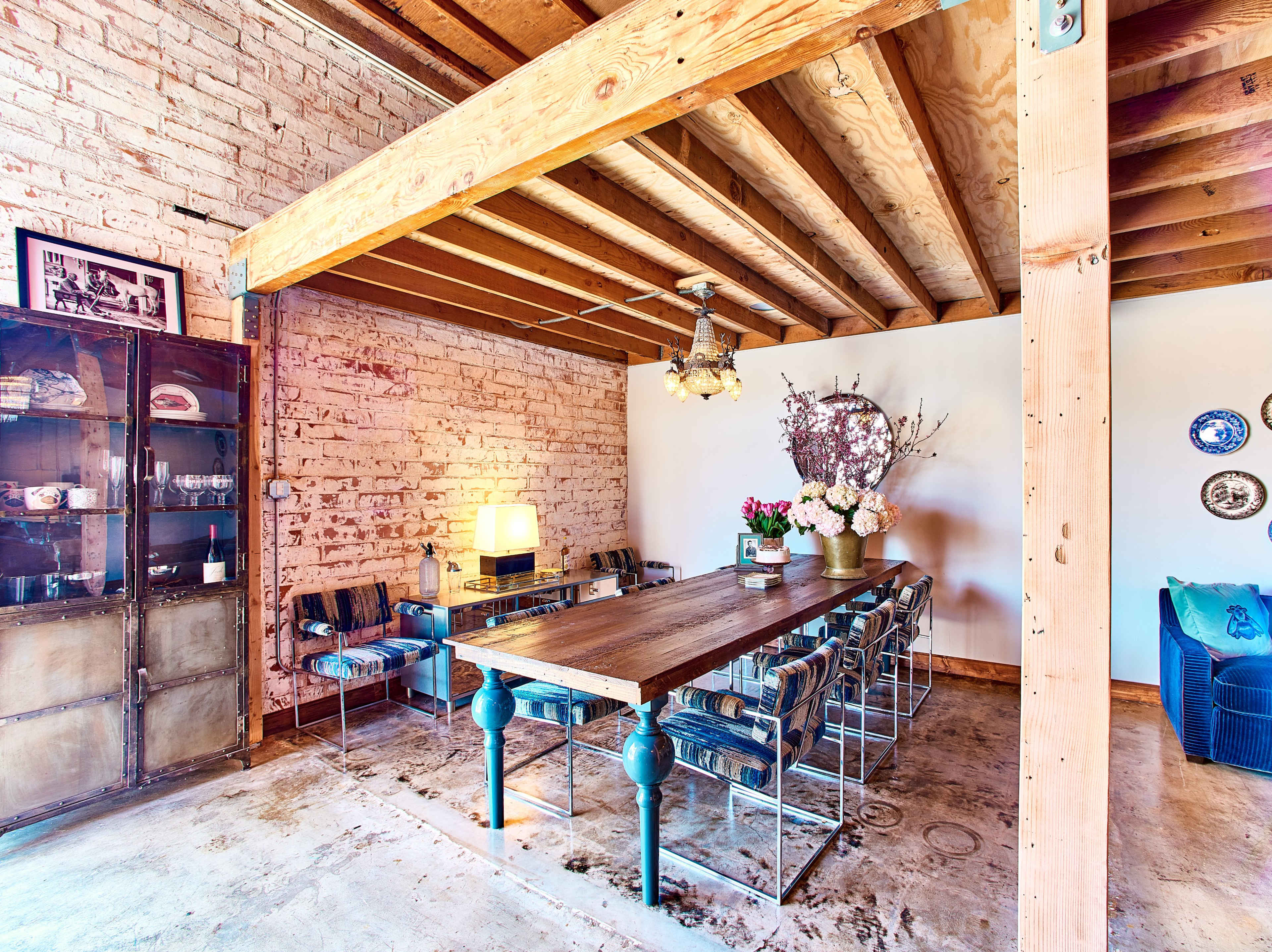The image shows a dining area with a long wooden table surrounded by metal chairs, featuring exposed brick walls and wooden beams overhead.