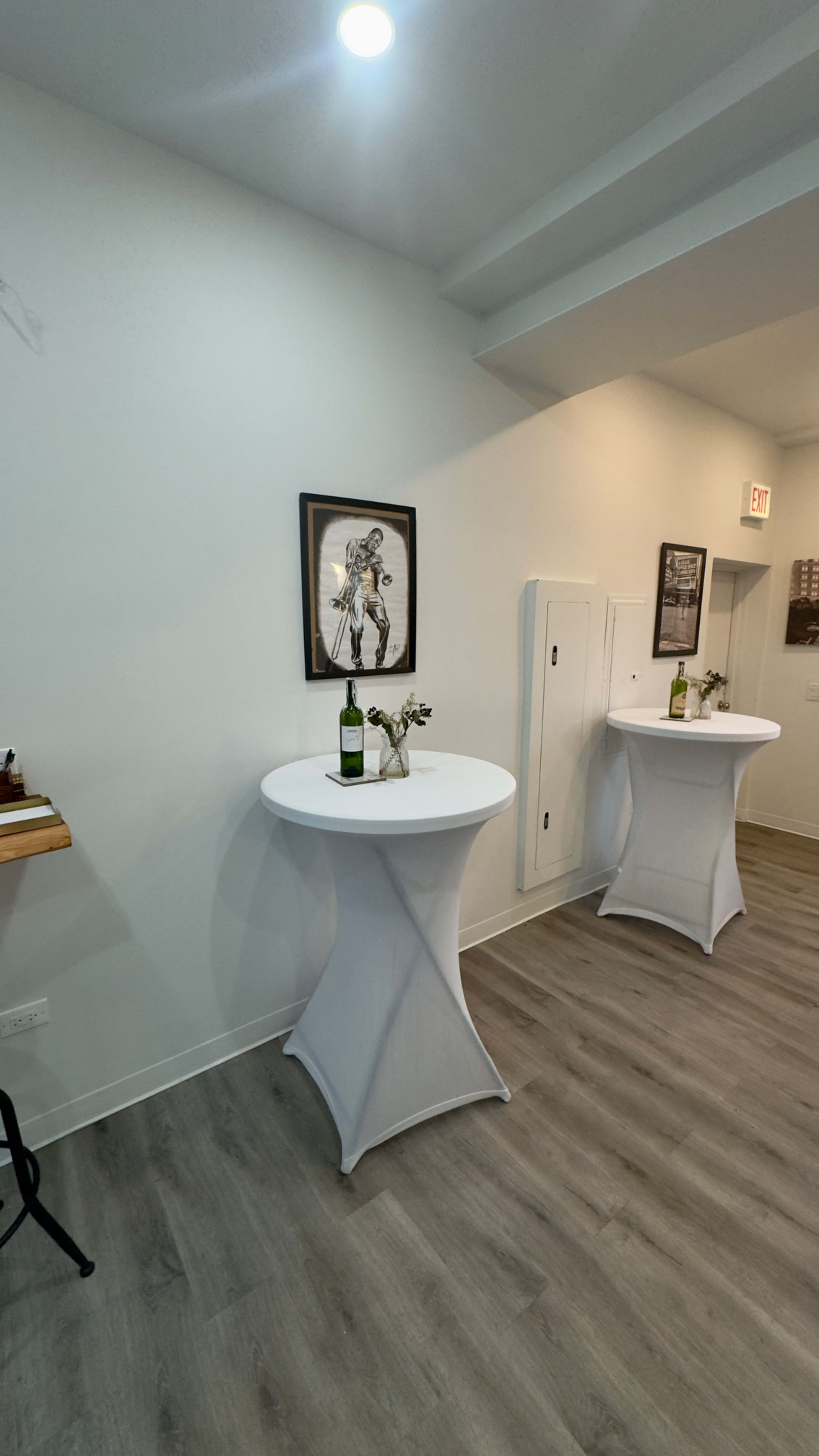 The image shows a bright room with two tall, white tables, each topped with a bottle of wine and a small arrangement of flowers, against a plain white wall featuring a framed artwork.