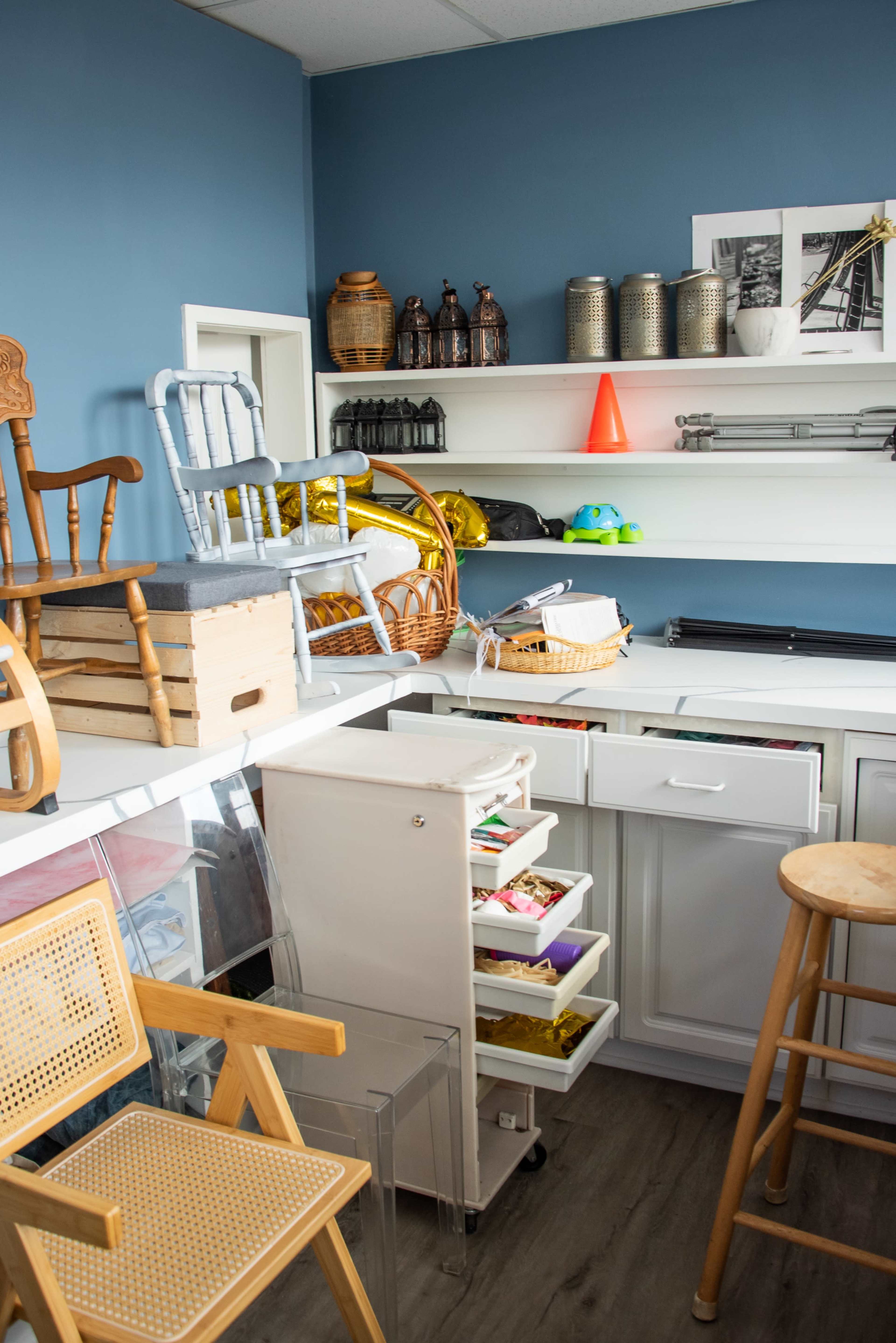 The image shows a cluttered room with wooden chairs, storage boxes, and various items organized on white shelves against a blue wall.