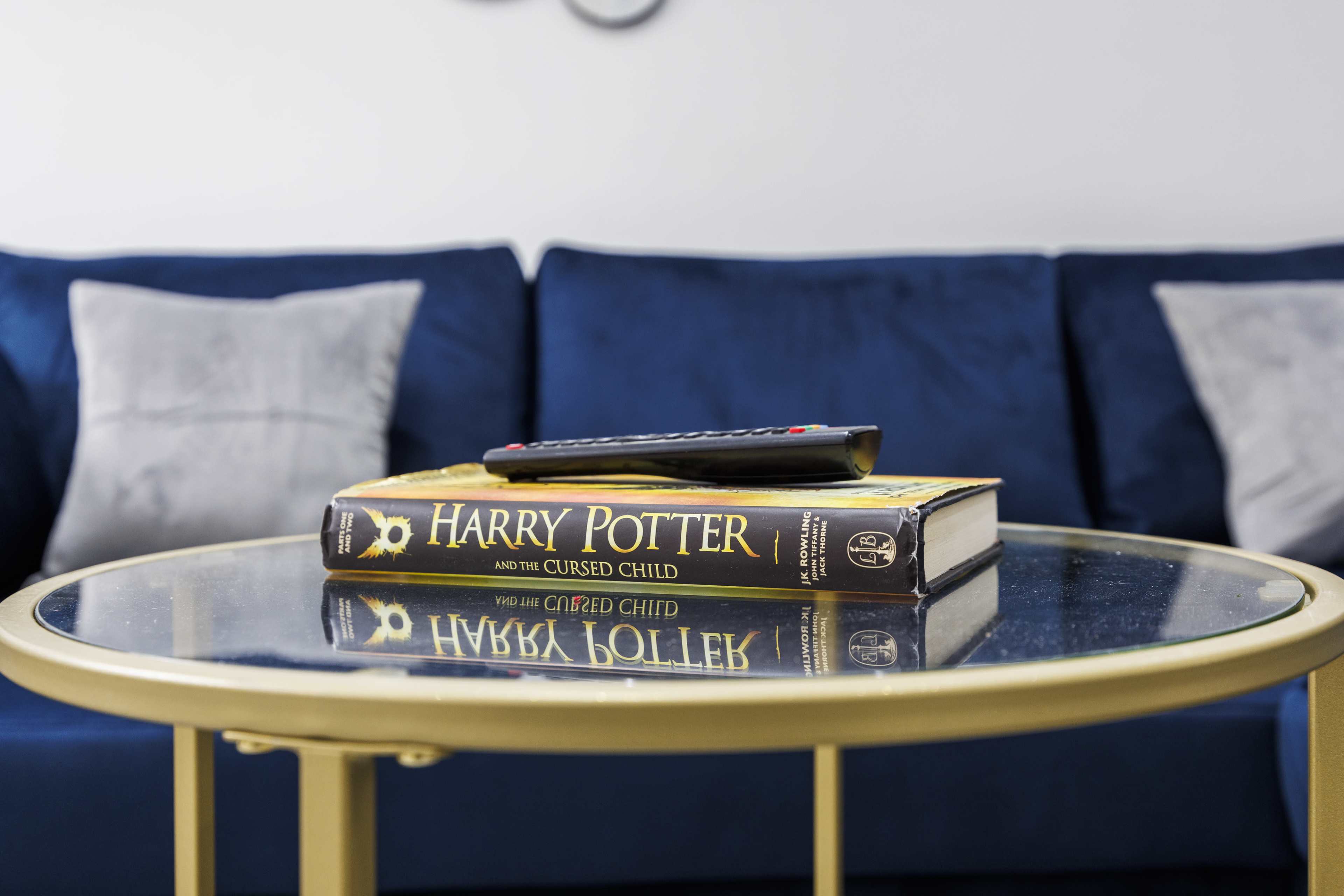 A copy of "Harry Potter and the Cursed Child" rests on a glass coffee table beside a remote control, with a blue couch in the background.