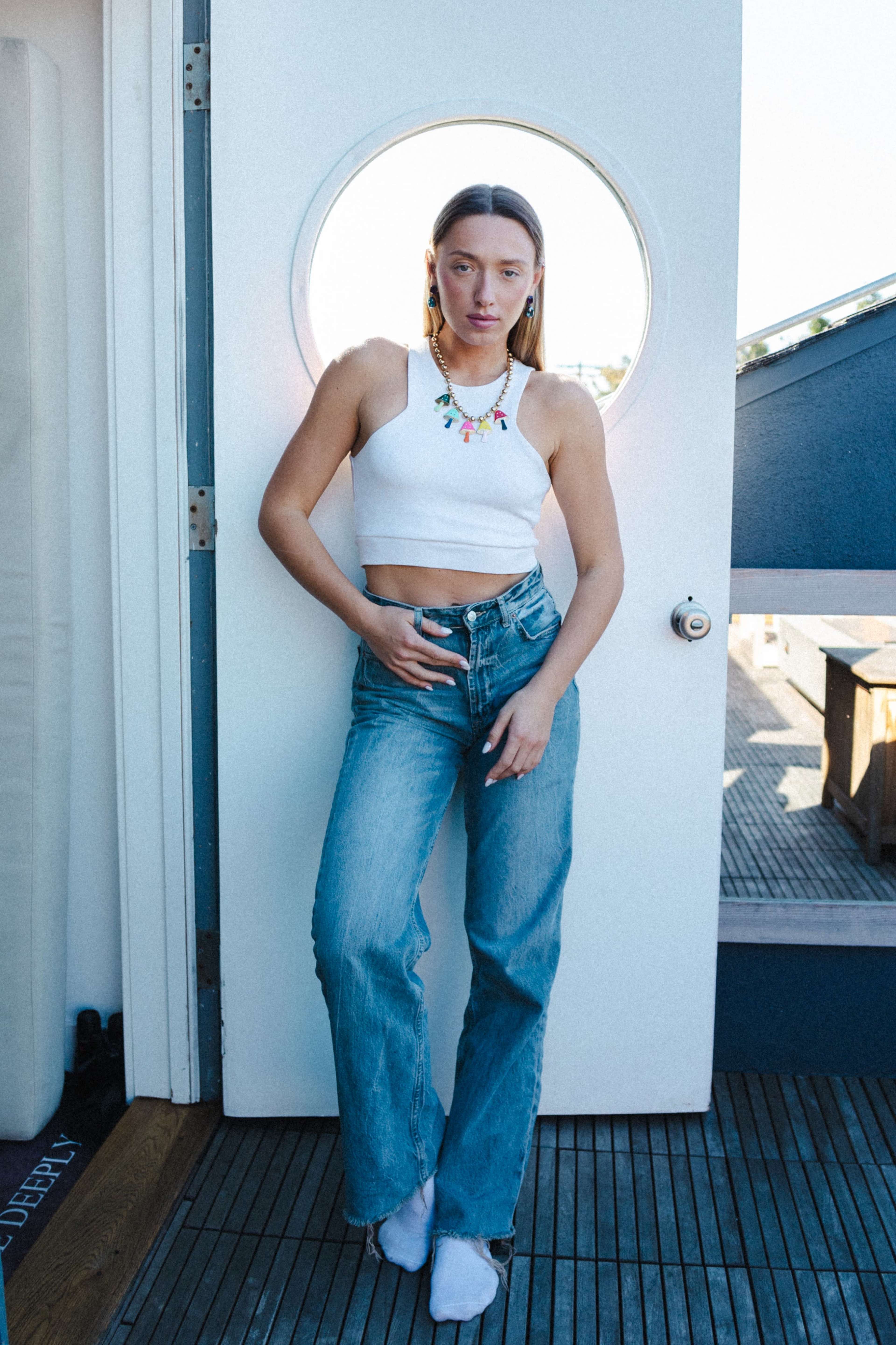 A person stands in front of a circular doorway, wearing a white crop top and blue jeans, with a colorful necklace.