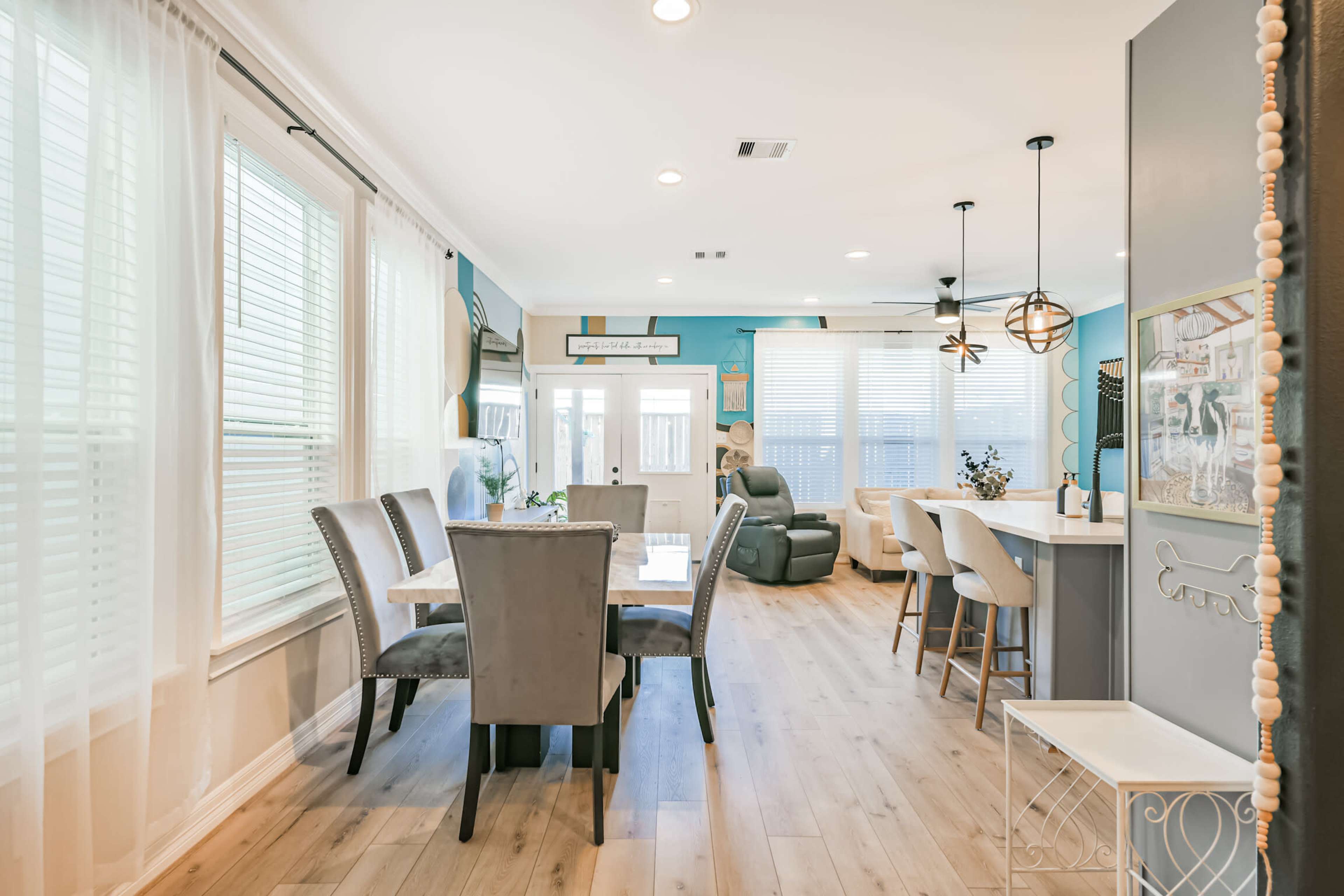 A dining area with gray upholstered chairs is adjacent to a kitchen bar and features large windows that allow natural light to illuminate the space.