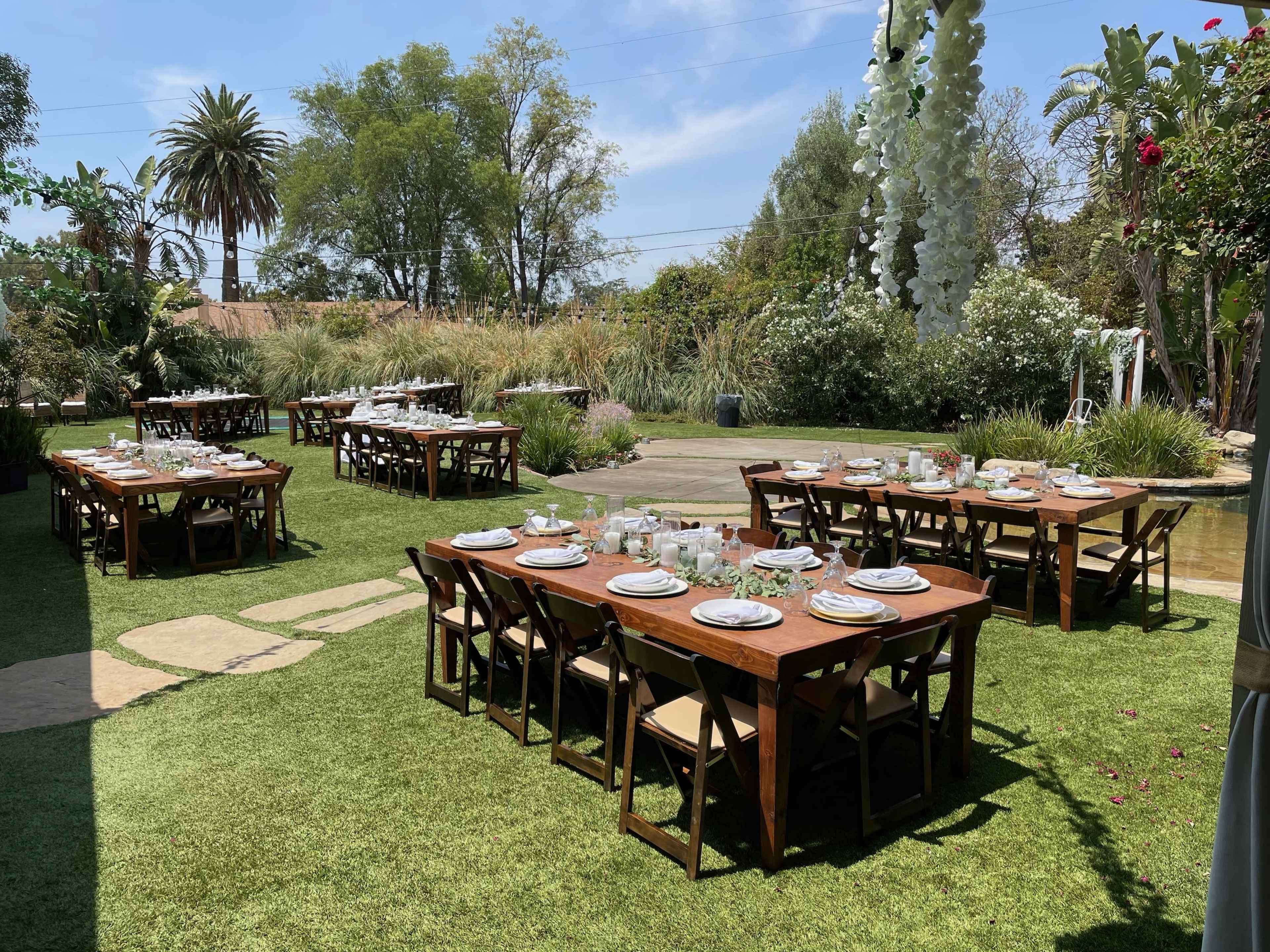 The image shows a setup for an outdoor dining event with multiple wooden tables arranged on a grass lawn, surrounded by greenery and palm trees.