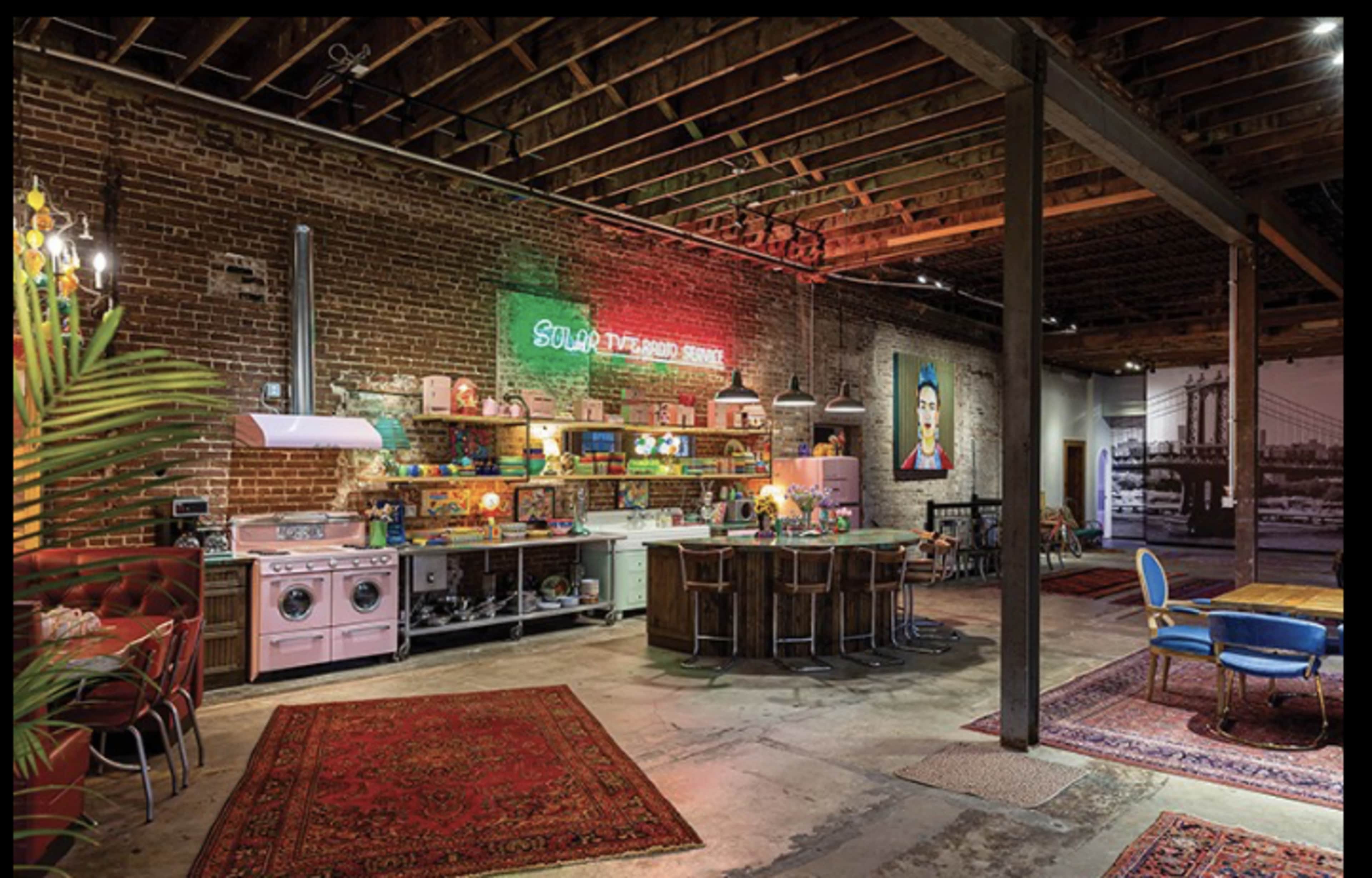 The image depicts a spacious industrial-style kitchen and living area with exposed brick walls, wooden beams, and a mix of colorful furnishings.