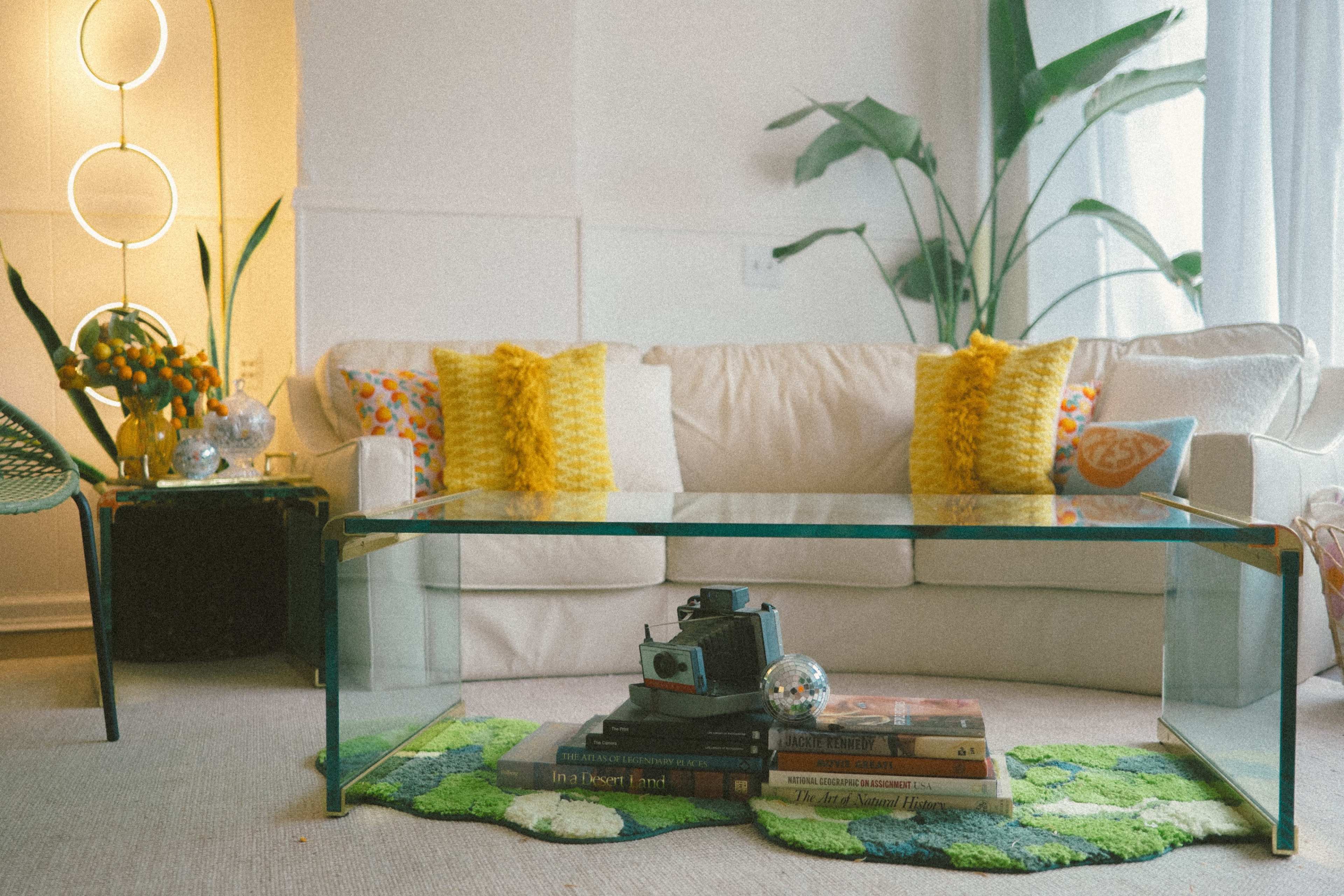 The scene features a cozy living room with a white sofa adorned with colorful pillows, a glass coffee table holding vintage books and a camera, and large green plants in the background.