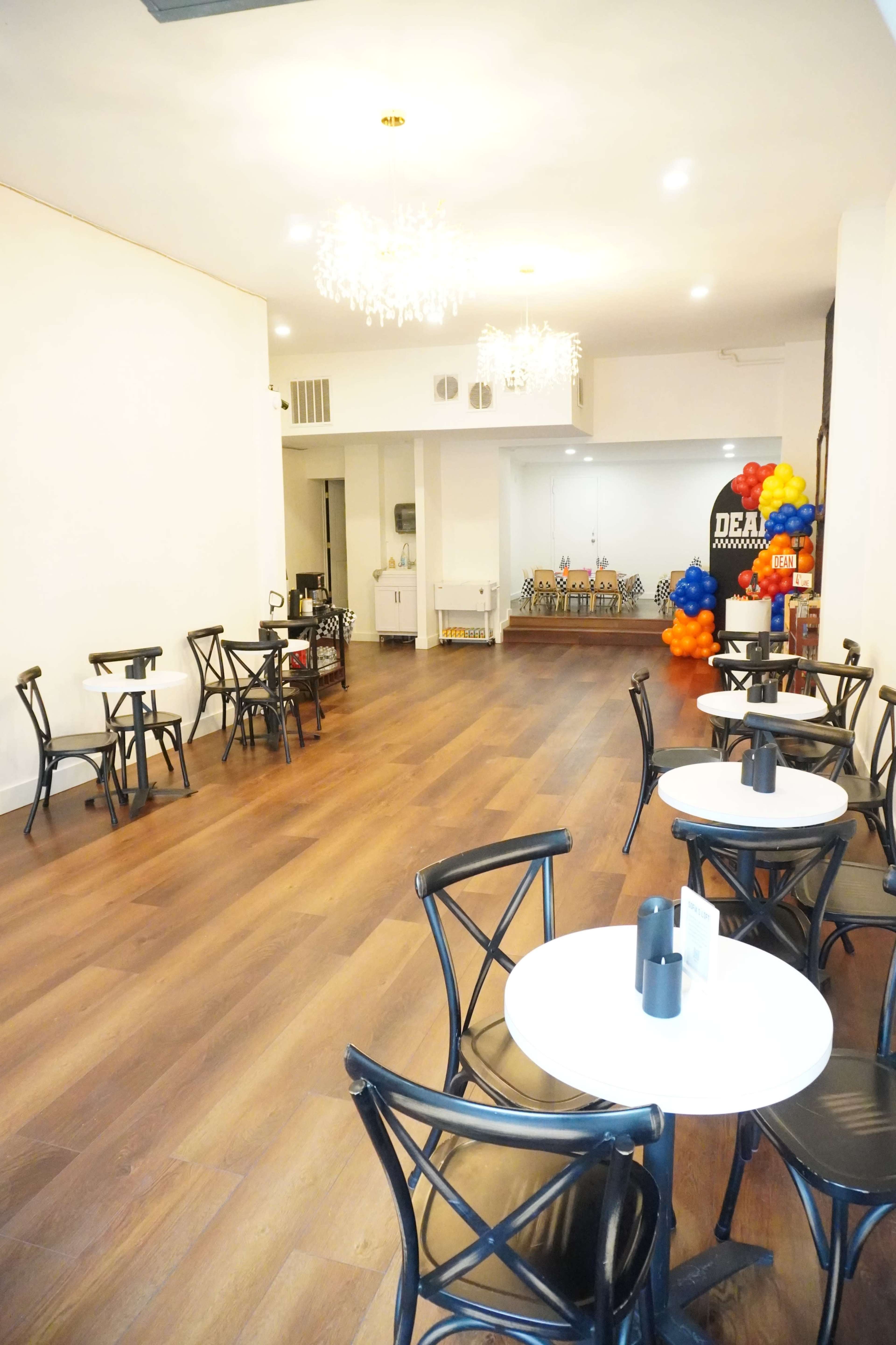 The image shows a spacious cafe with black wooden chairs and small round tables arranged on polished wood flooring, featuring a decorative backdrop with colorful balloons in one corner.