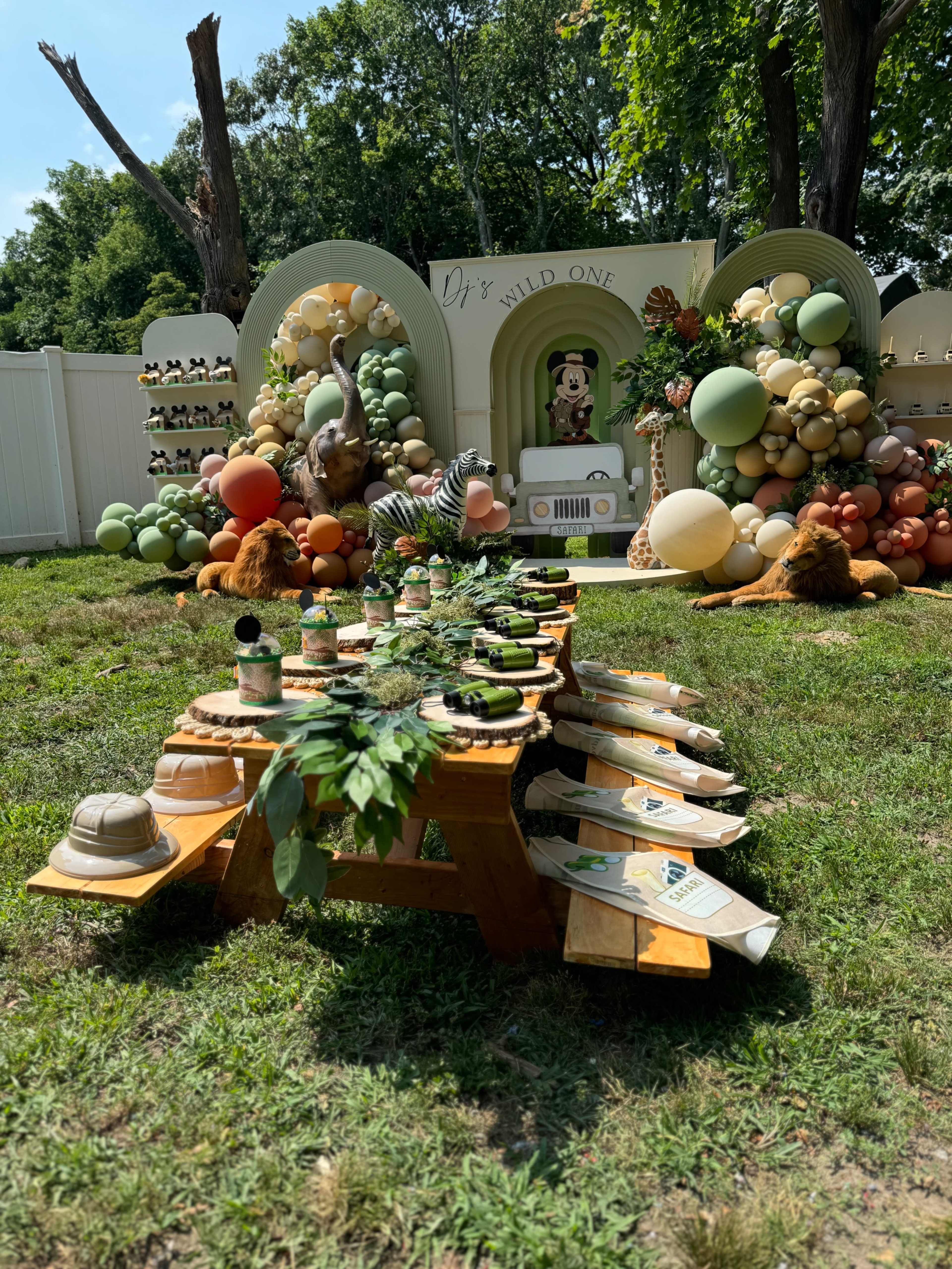 The image shows a colorful outdoor party setup featuring a table adorned with plants and greenery, surrounded by elaborate decorations of balloons and animal figures.