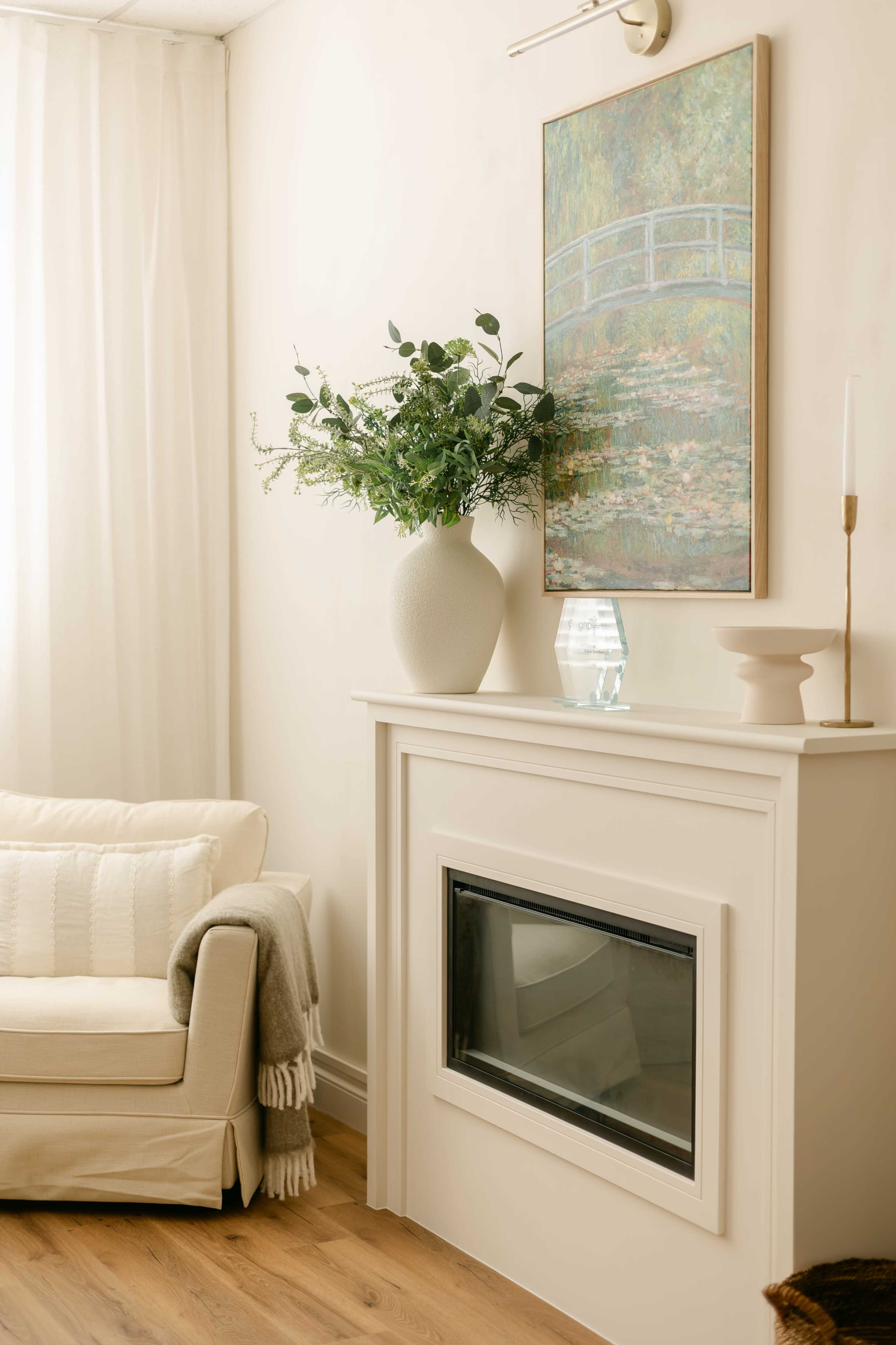 A cozy living room features a neutral-colored sofa, a fireplace with a modern mantle, and a large vase of greenery next to a framed landscape painting.