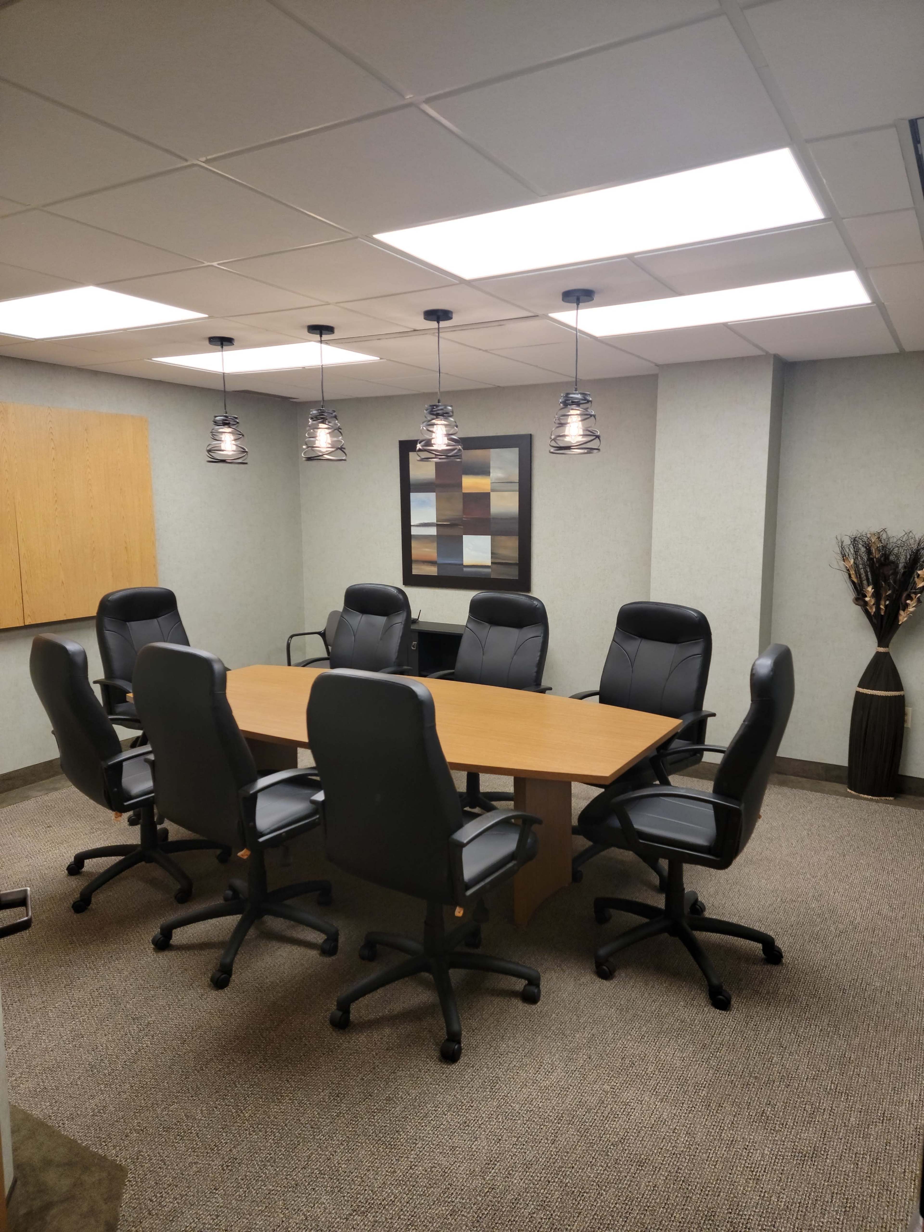 A modern conference room features a large wooden table surrounded by six black office chairs and decorative lighting.