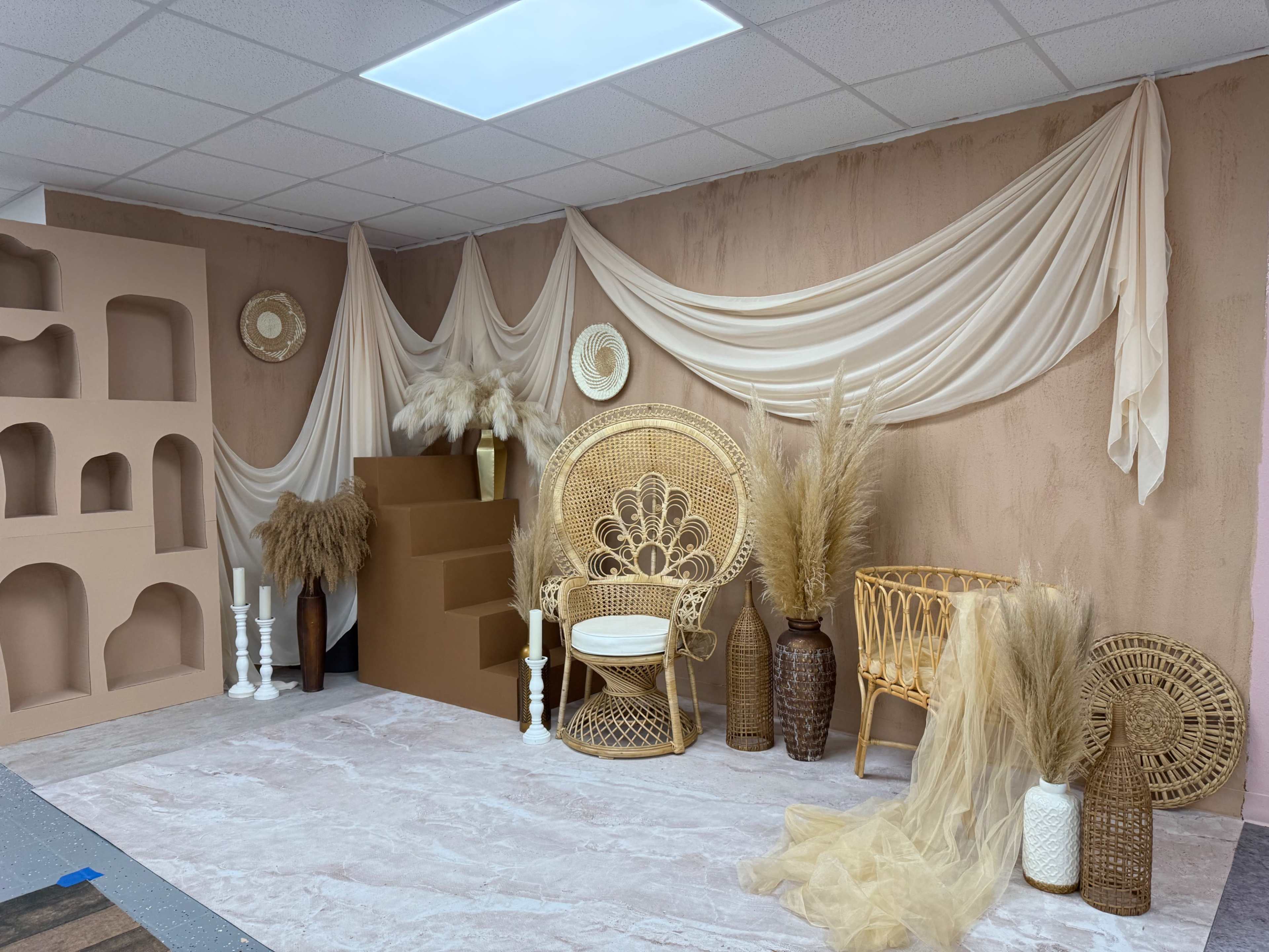 The image shows a styled indoor space featuring neutral-colored drapery, rattan furniture, decorative vases, and a staircase against a textured wall.