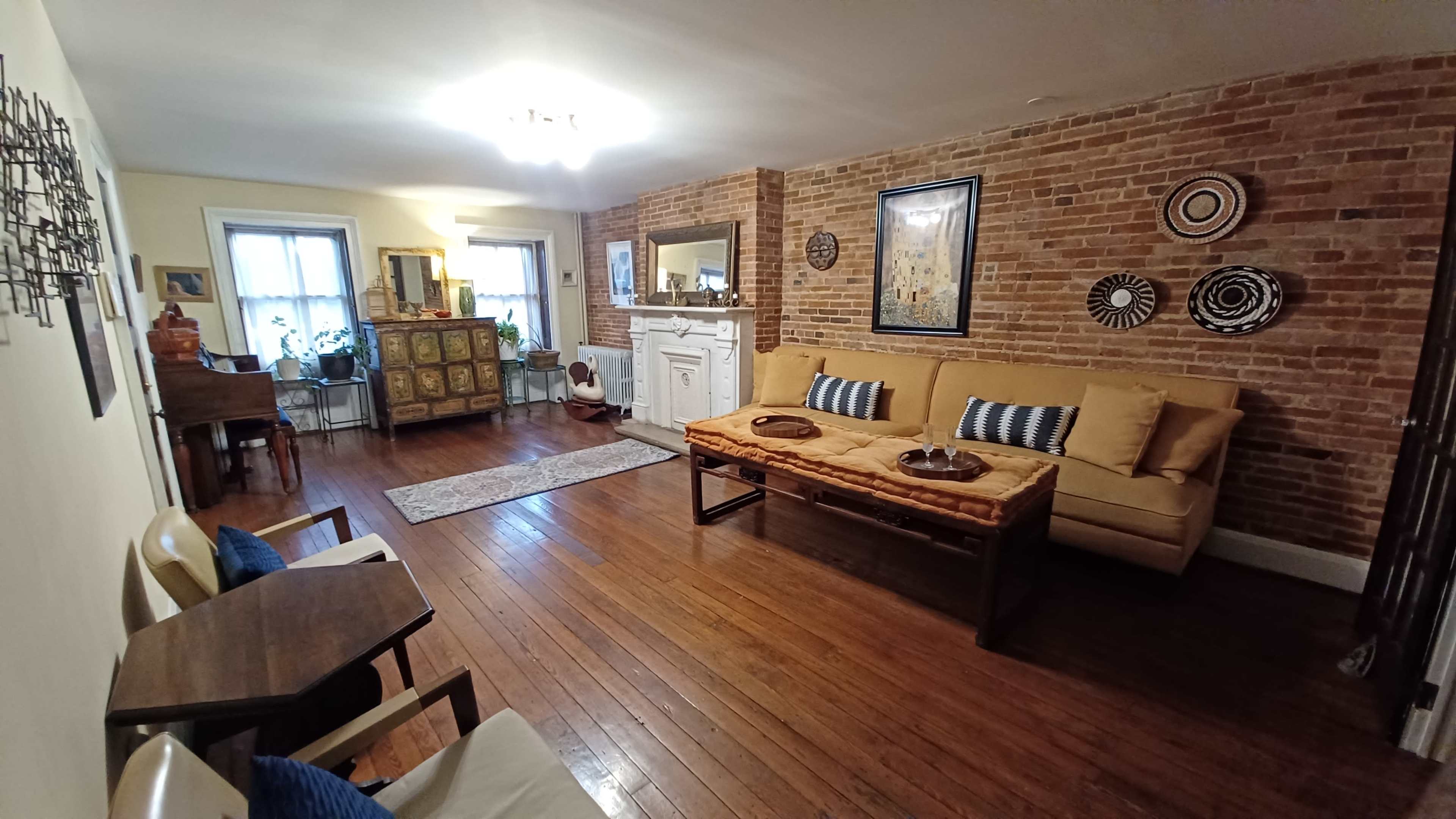 A living room with exposed brick walls, wooden flooring, and a sofa with decorative cushions, along with a dining area and various pieces of furniture.