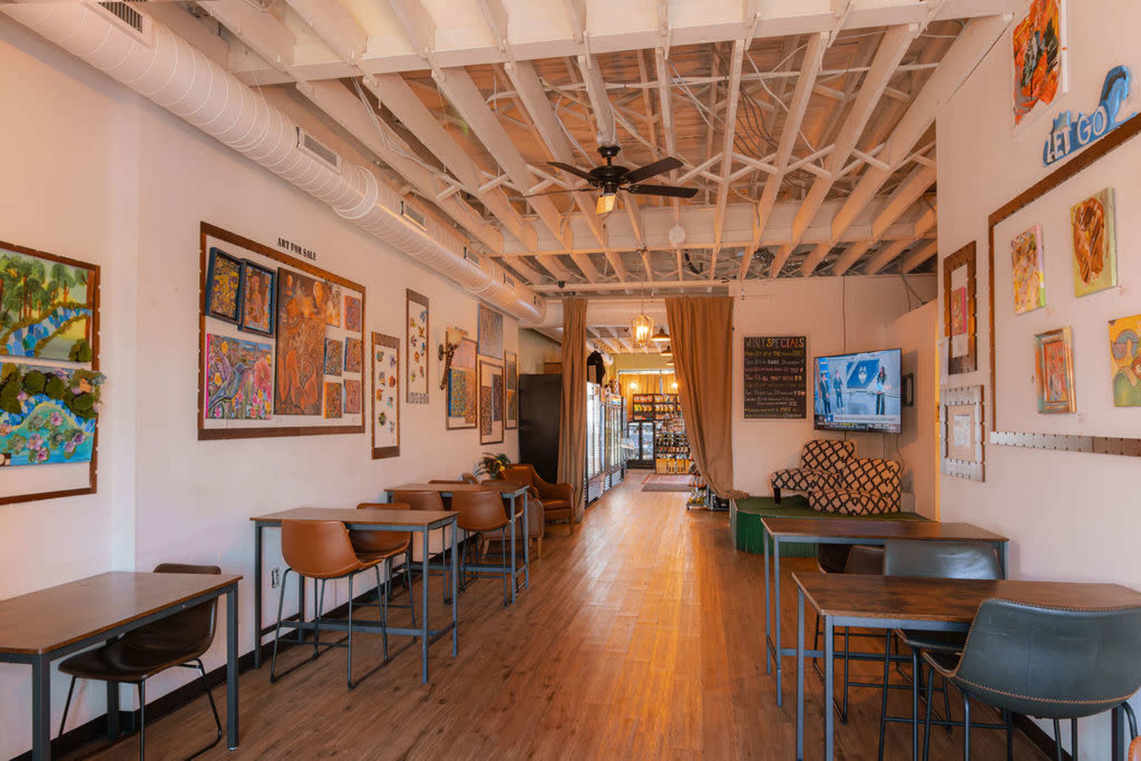 The image shows the interior of a cafe with wooden flooring, several tables and chairs, and walls adorned with colorful artwork.