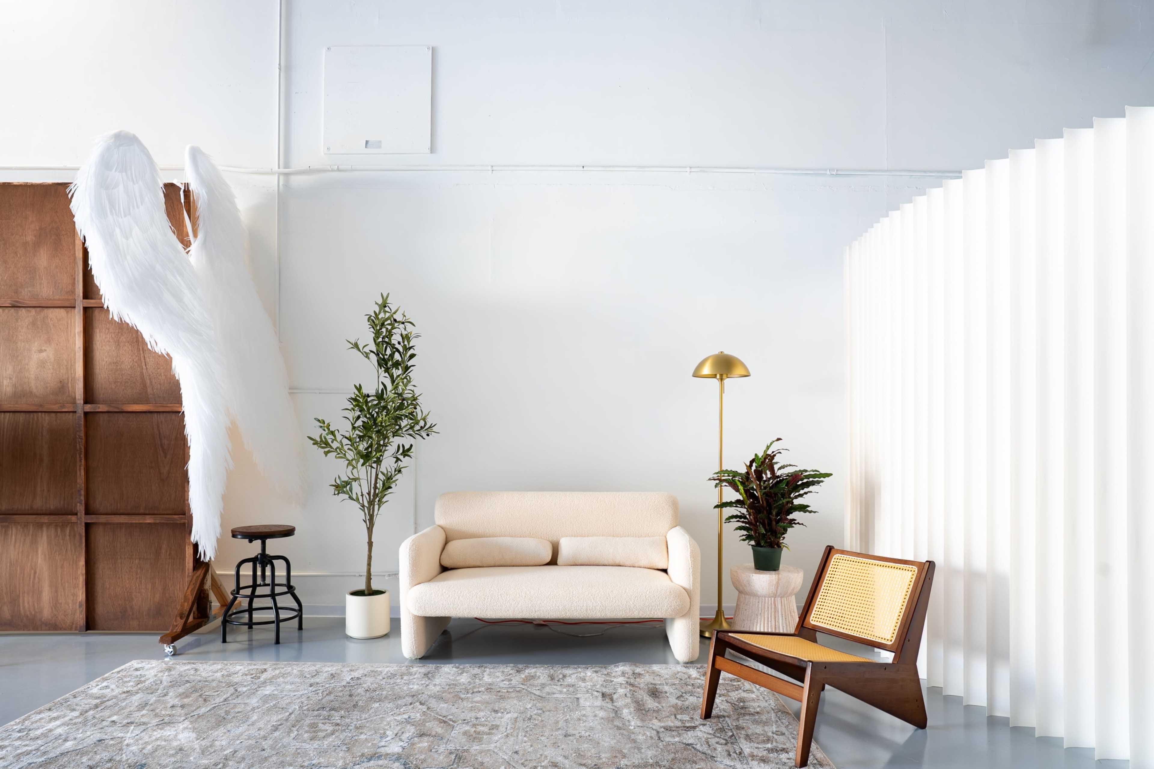 The image shows a minimalist interior with a light-colored sofa, a wooden chair, potted plants, and a large decorative angel wings installation against a white wall.