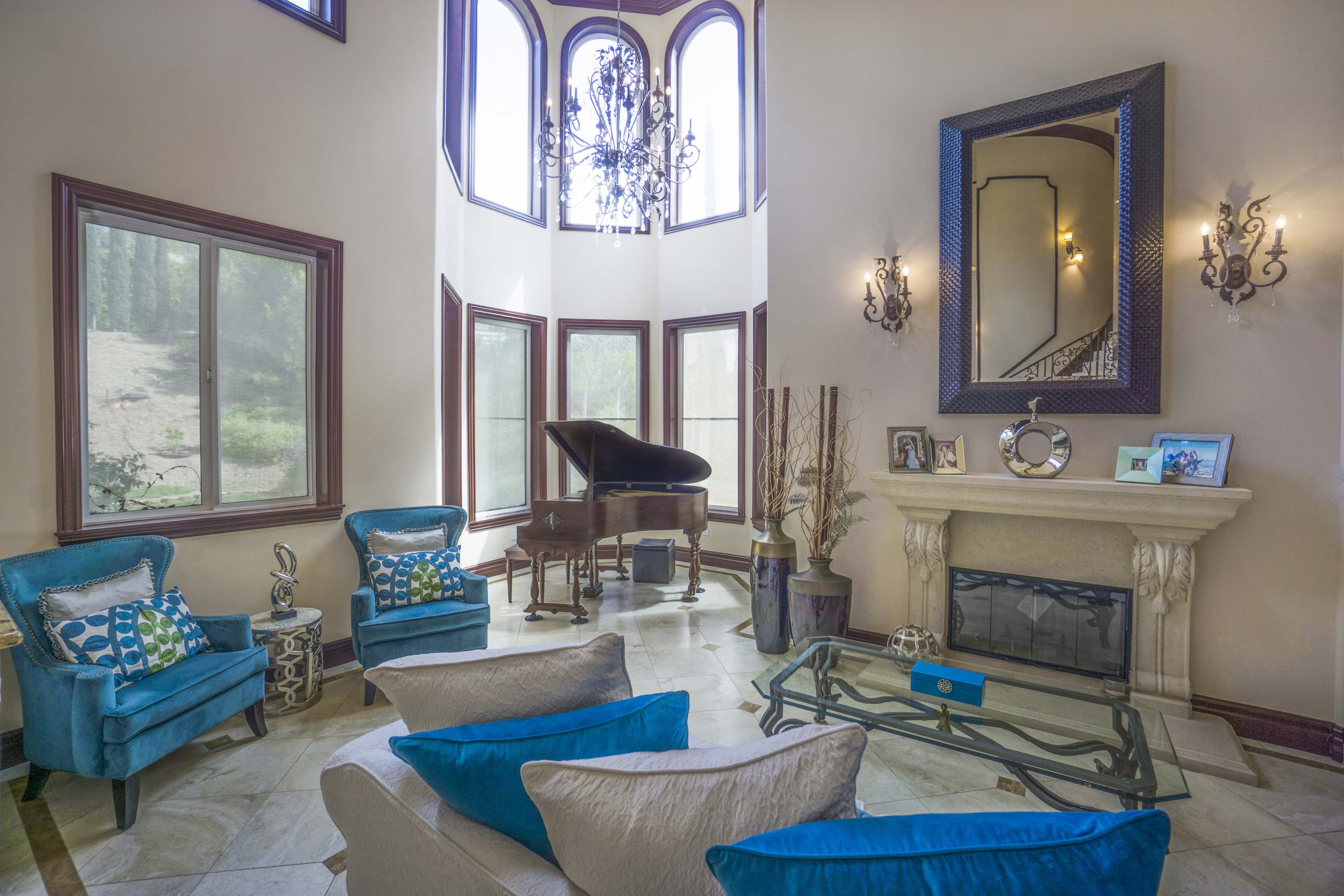 A spacious living room features a grand piano near large windows, with two blue chairs and a glass coffee table in front of a fireplace.