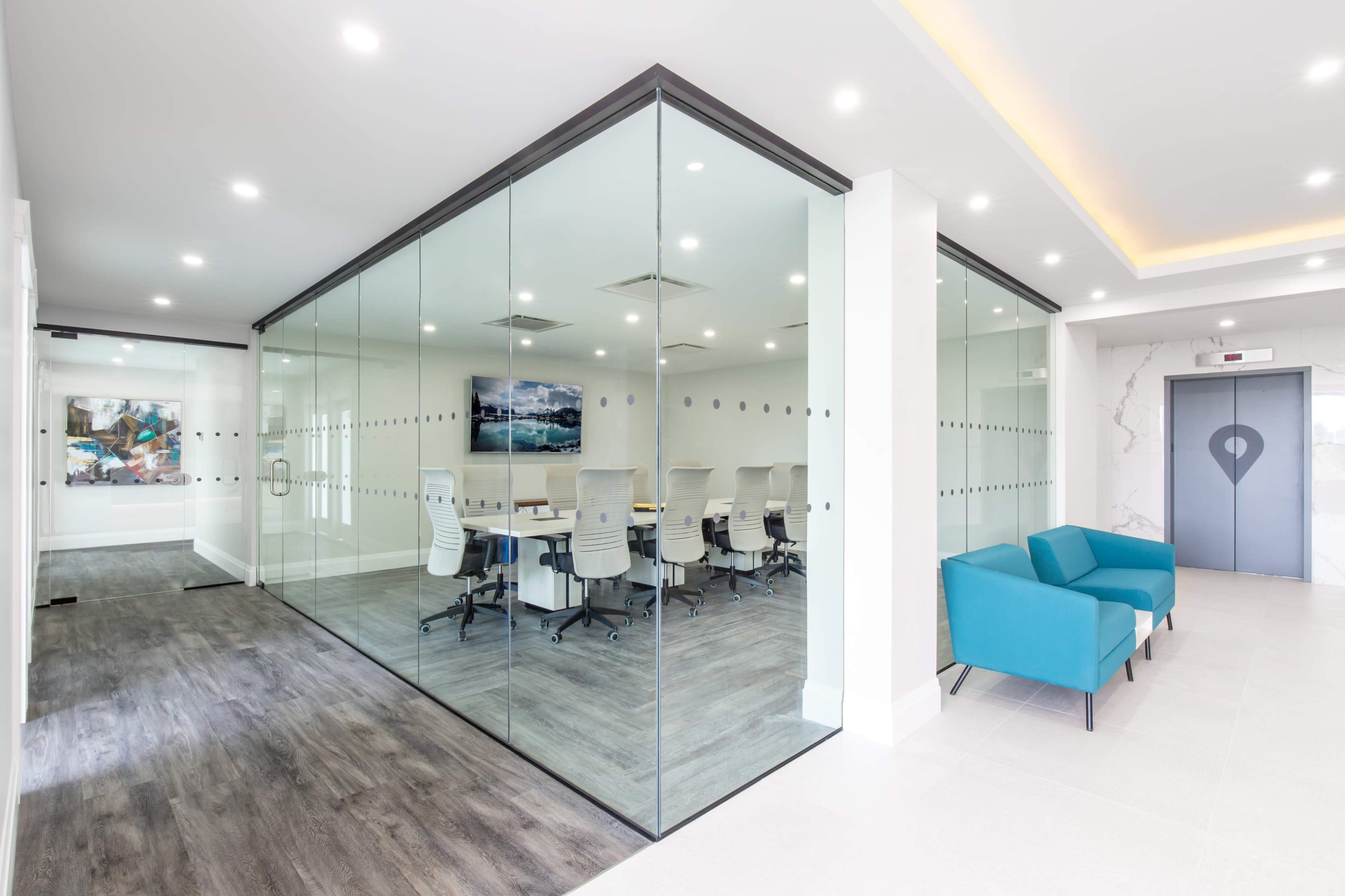 A modern office space featuring a glass-walled meeting room with several white chairs and a table, alongside a seating area with turquoise chairs.
