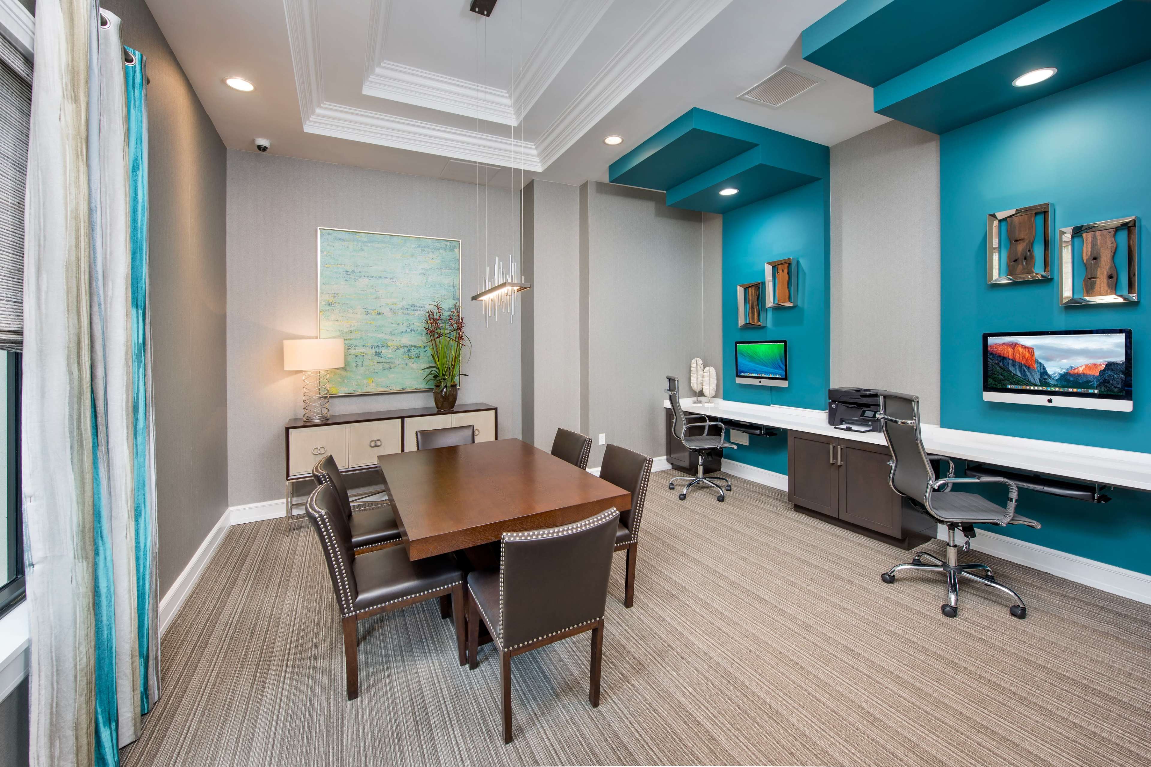 A modern office space featuring a wooden dining table, dark chairs, a light fixture, and two workstations against teal walls, with artwork and decorative elements displayed.