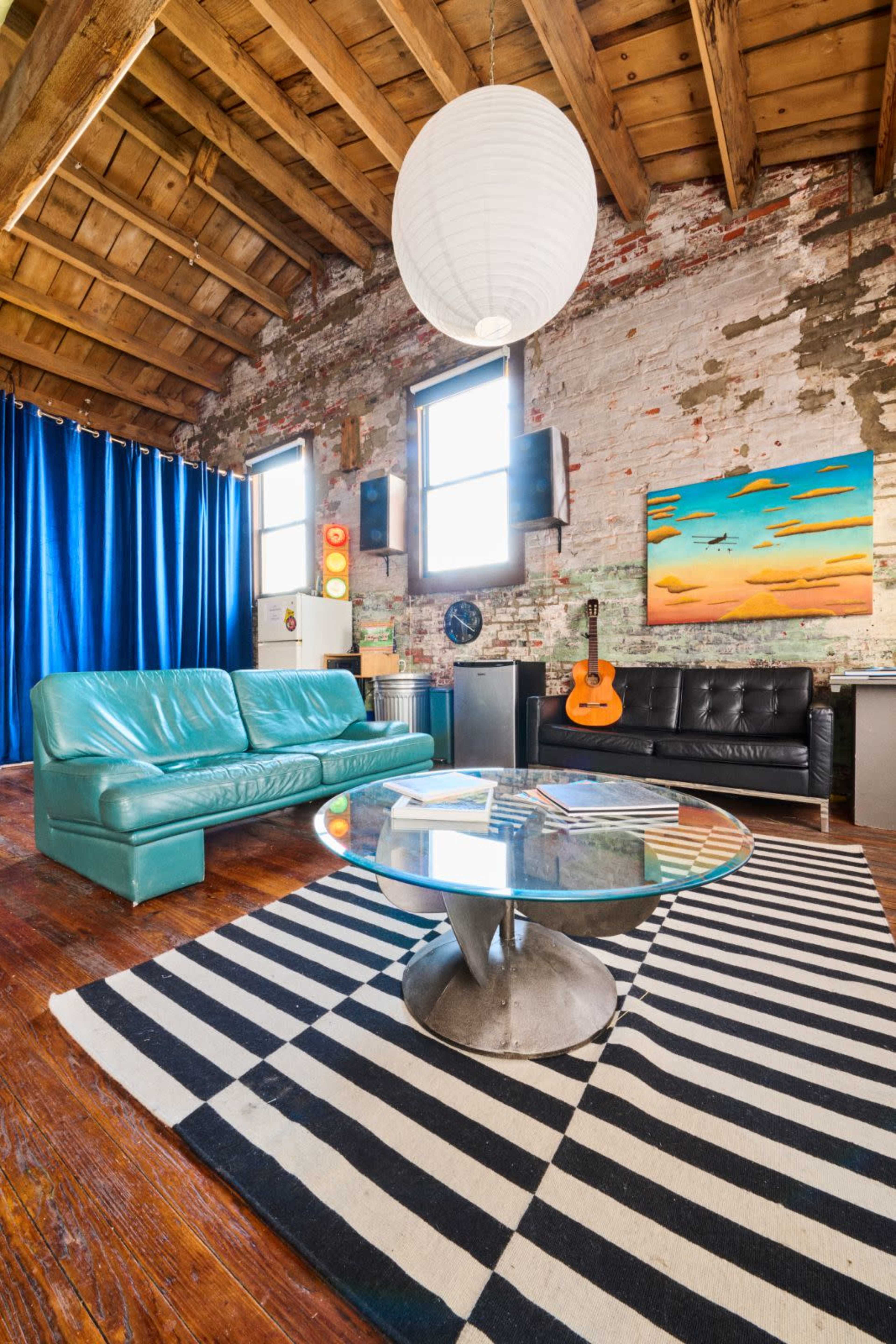 A spacious living room featuring two couches, a round glass table on a striped rug, an acoustic guitar, and large windows with blue curtains.