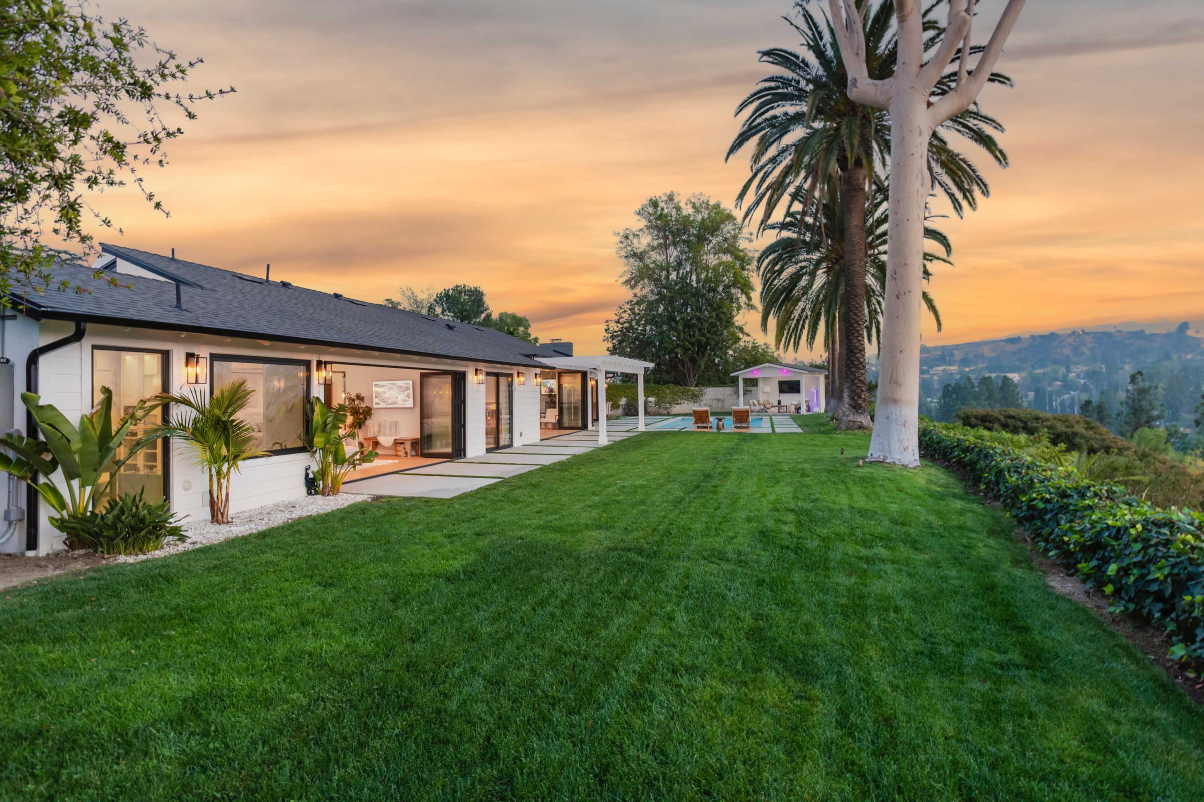 A modern house with a spacious lawn, palm trees, and a sunset in the background.