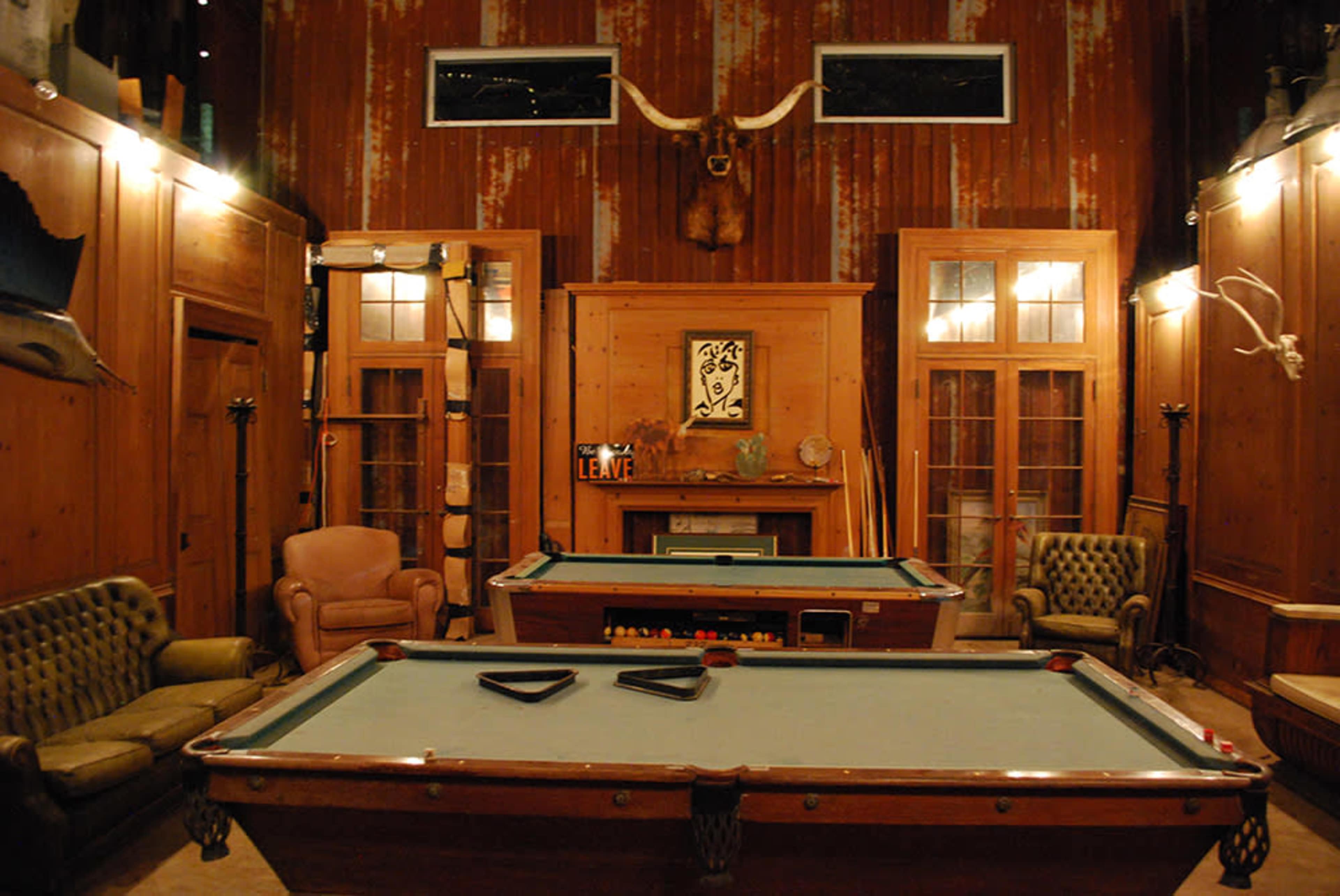 A rustic room with two pool tables, wooden walls, and a cozy seating area featuring vintage furniture.