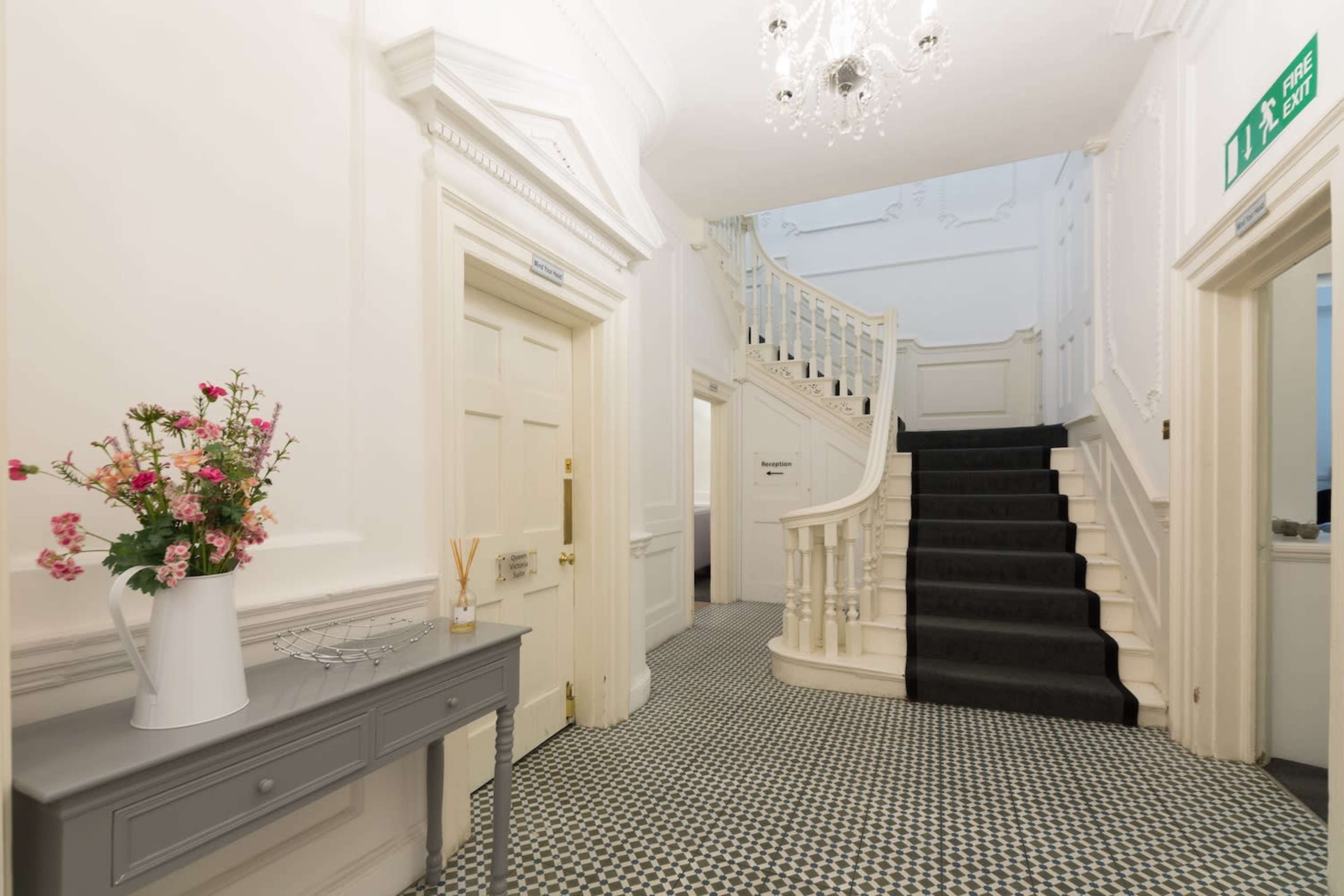 The image shows a bright hallway with a staircase, a small table with a vase of flowers, and patterned flooring.
